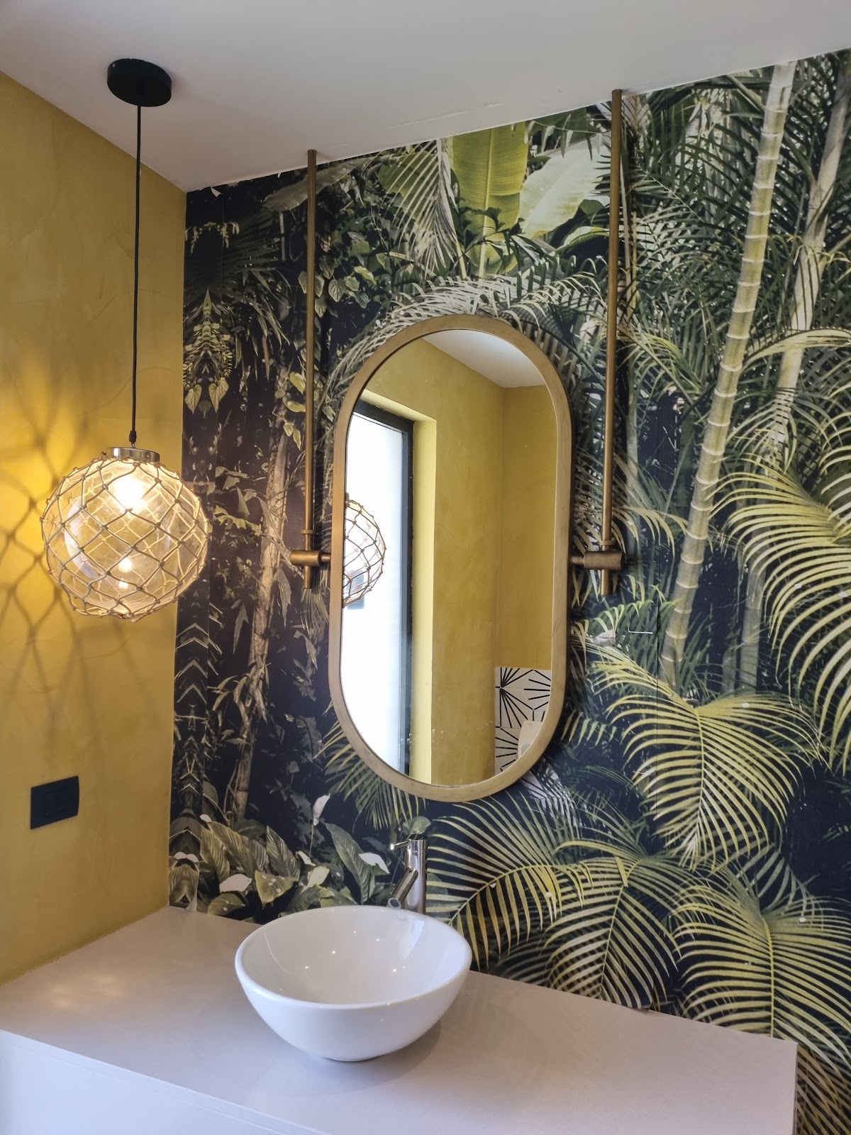 Bathroom vanity with white bowl sink, oval mirror, and tropical palm-leaf wallpaper in navy and gold tones.