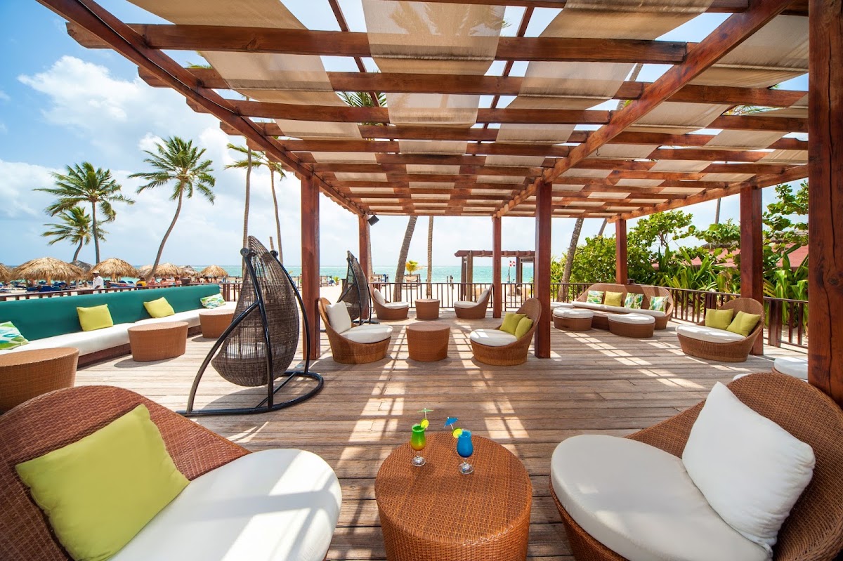 Wooden pergola lounge area with wicker chairs, lime cushions, and hanging egg chair overlooking pool and ocean at Punta Cana Princess.