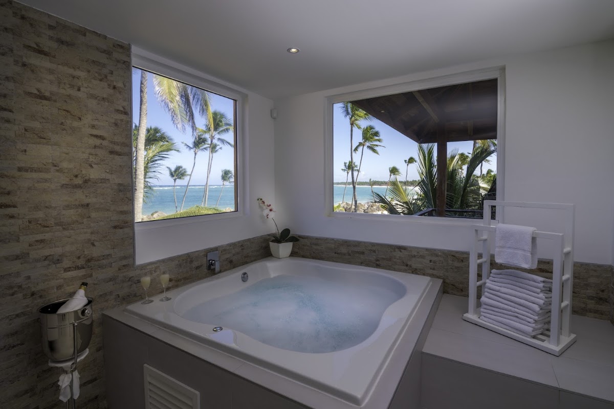 Spa bathtub with ocean and palm tree views through large windows in a modern bathroom at Punta Cana Princess.