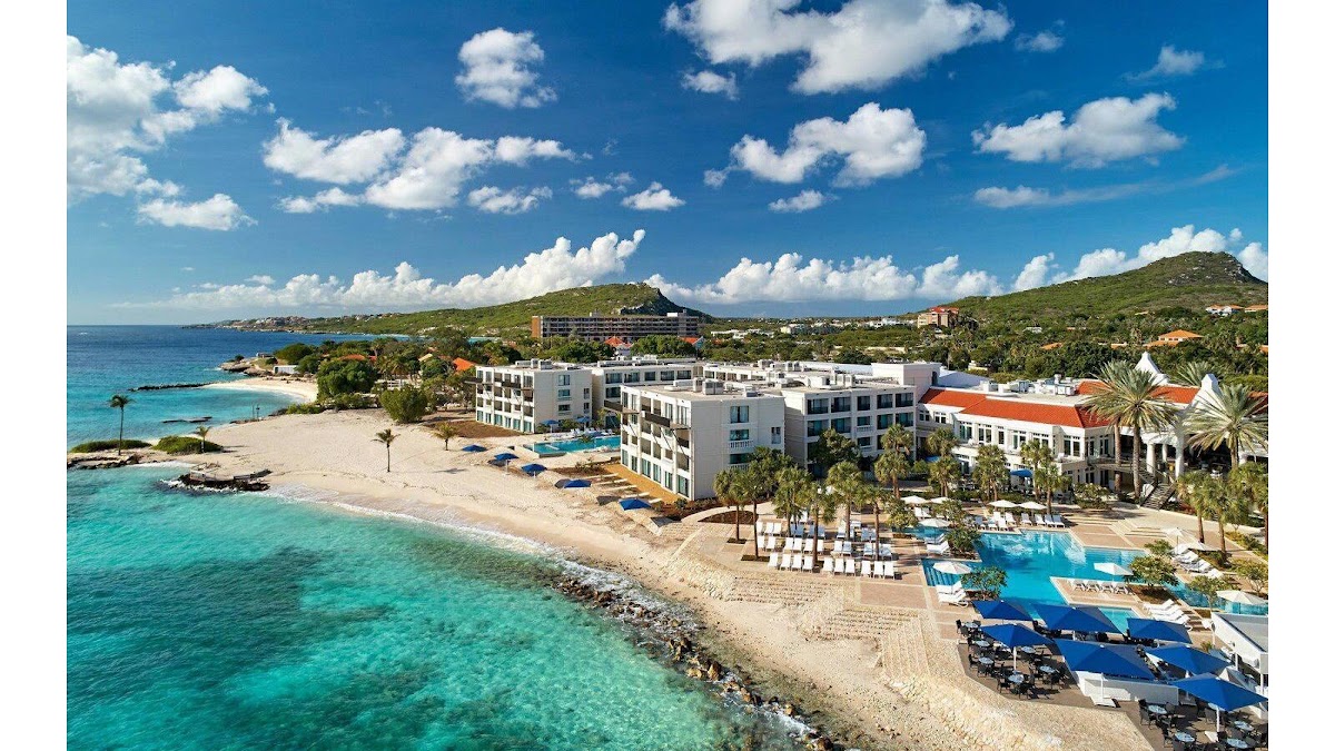 White multi-story resort buildings with blue pool and lounge chairs sit directly on a sandy beach beside turquoise Caribbean water, with green hillsides and palm trees in the background.