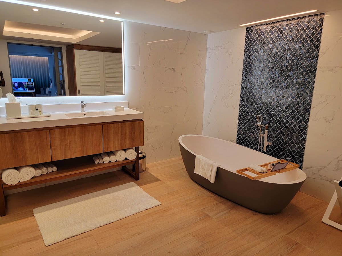 Modern bathroom with dark freestanding tub, marble-look walls, wood vanity with rolled towels, and geometric tile shower surround.