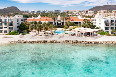 The Pyrmont Curaçao beachfront with turquoise water, white buildings, red-roofed central structure, palm trees, and a rocky shoreline under blue sky.