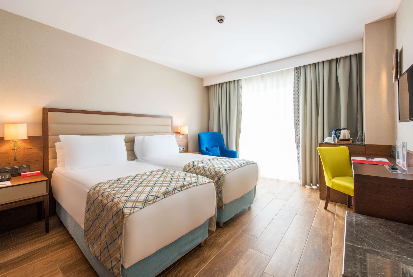 Resort guest room with two twin beds, wood furnishings, blue and yellow accent chairs, and balcony access at Ramada Plaza by Wyndham Antalya.