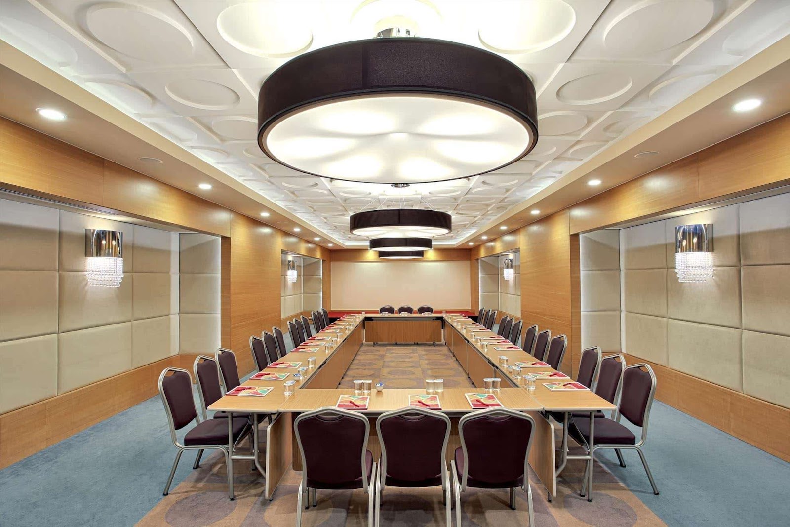 Modern conference room with long wooden table, burgundy chairs, cream padded walls, and circular black pendant lights from ceiling.