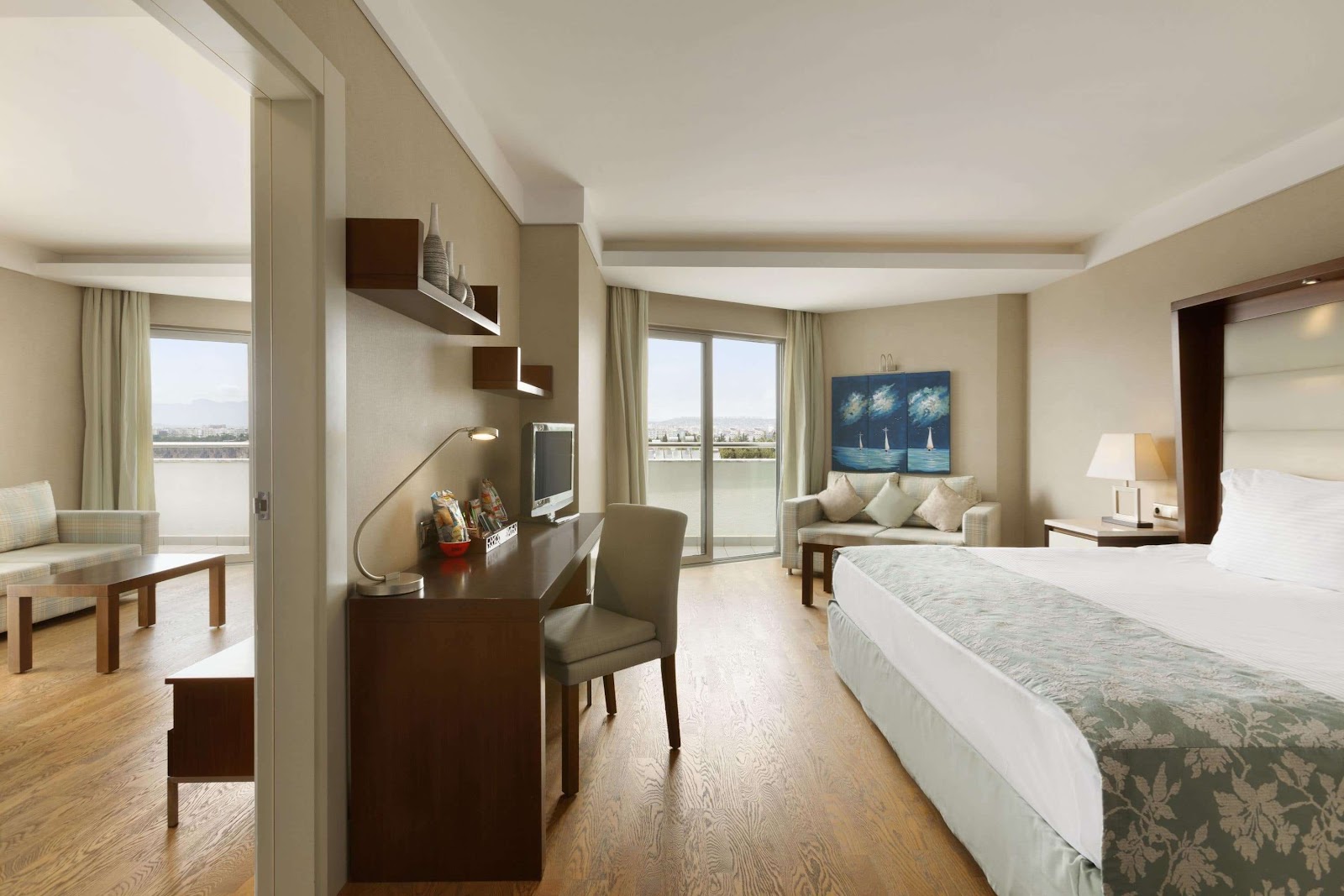 Spacious resort suite with queen bed, work desk, and seating area, floor-to-ceiling windows overlooking water and mountains.