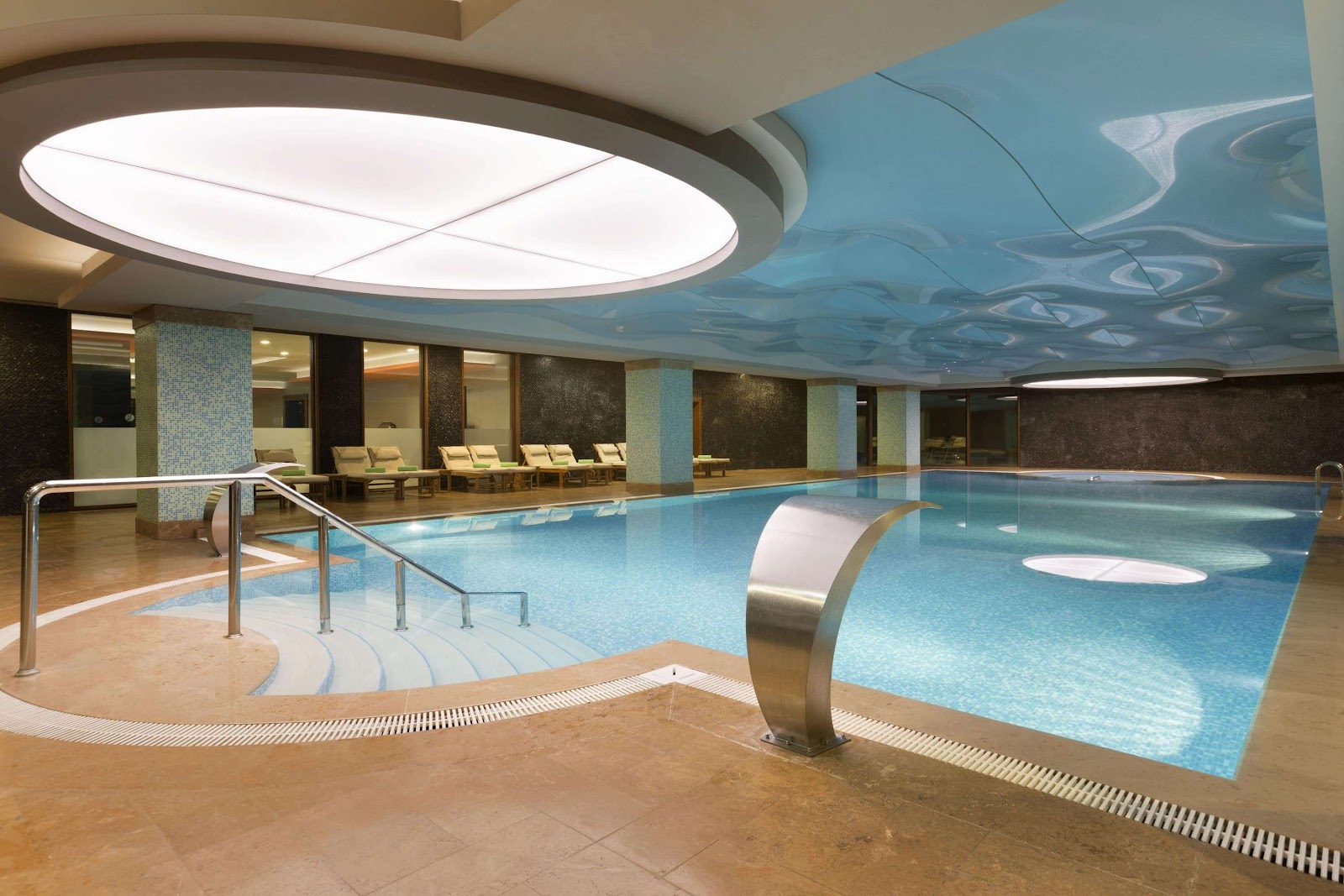 Indoor swimming pool with blue water, metal railings, lounge chairs along the side, and illuminated ceiling with decorative pattern.