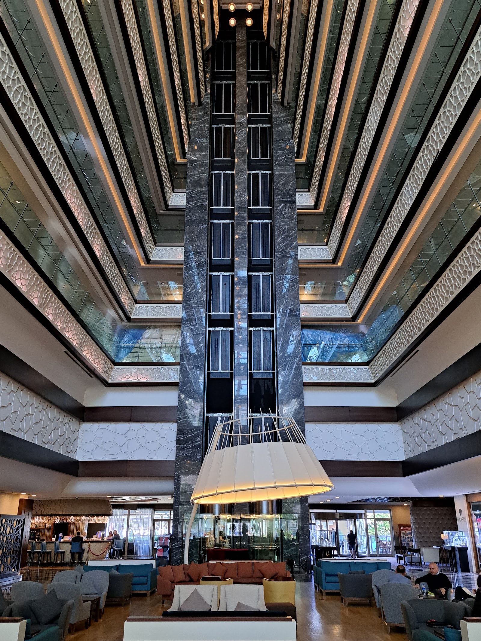 Soaring atrium with symmetrical dark marble columns, geometric patterned walls, and a large pendant light fixture over seating areas below at Ramada Plaza by Wyndham Antalya.