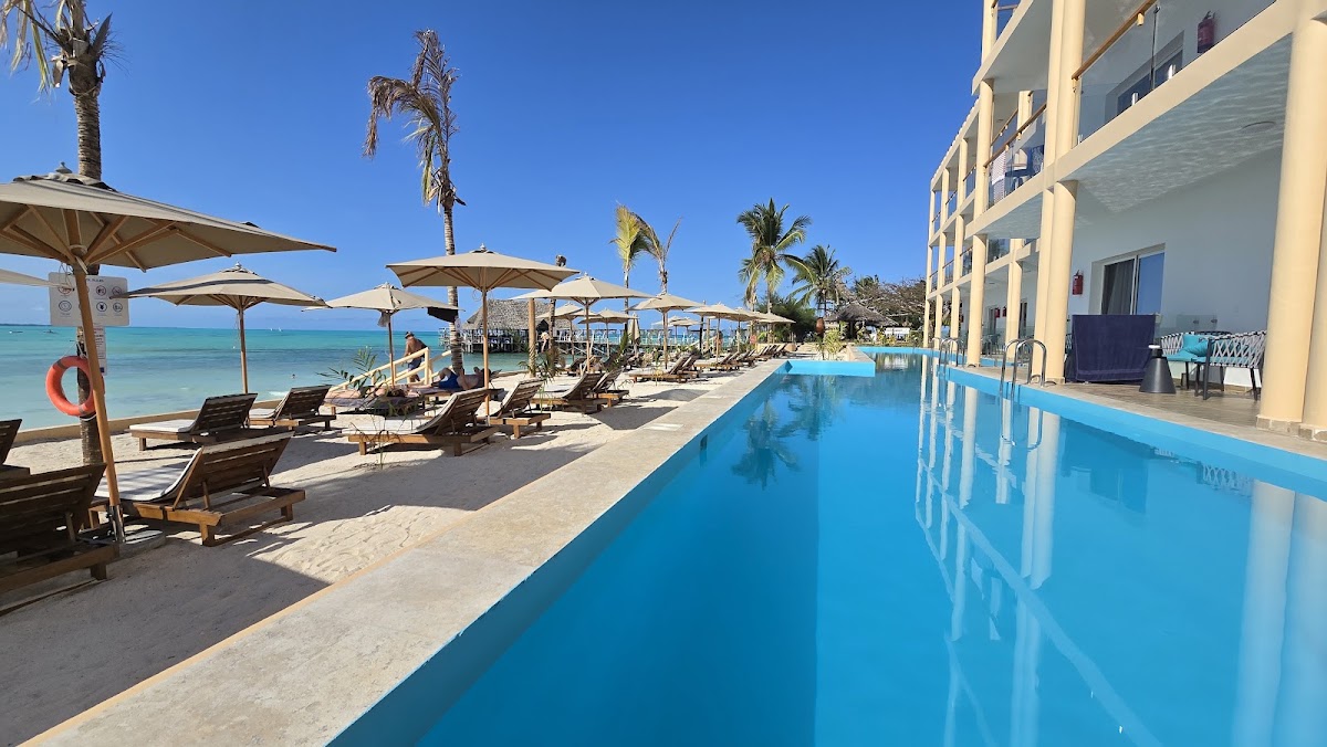 Blue swimming pool and cream-colored building with lounge chairs, umbrellas, and beach beyond under clear sky at Reef & Beach Resort.