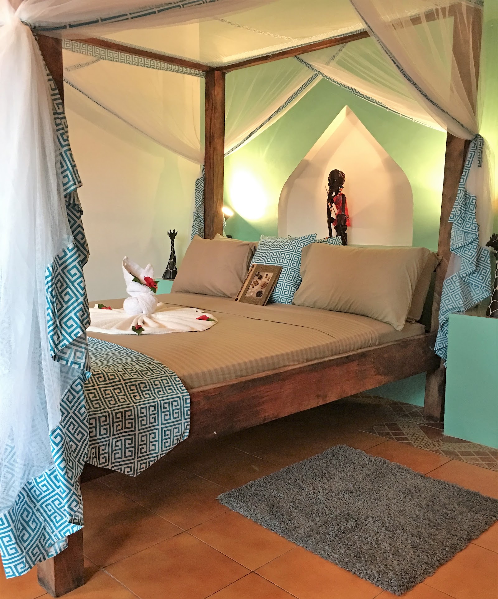 Four-poster bed with sheer curtains and geometric patterned drapes in a room with green-lit archway and terra cotta tile floor at Reef & Beach Resort.