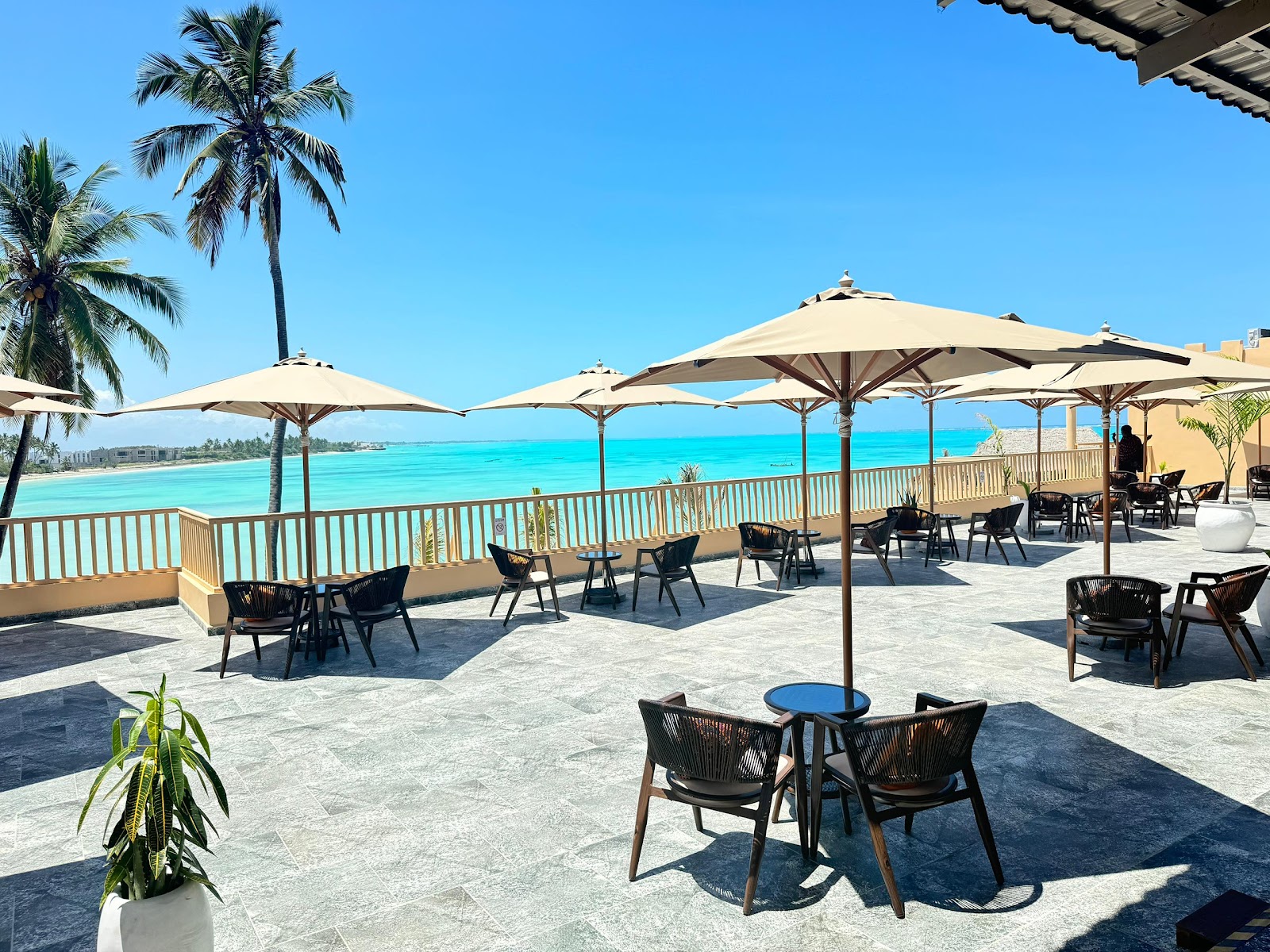 Shaded seating area on a waterfront terrace overlooking turquoise ocean at Reef & Beach Resort, with palm trees and clear blue sky.