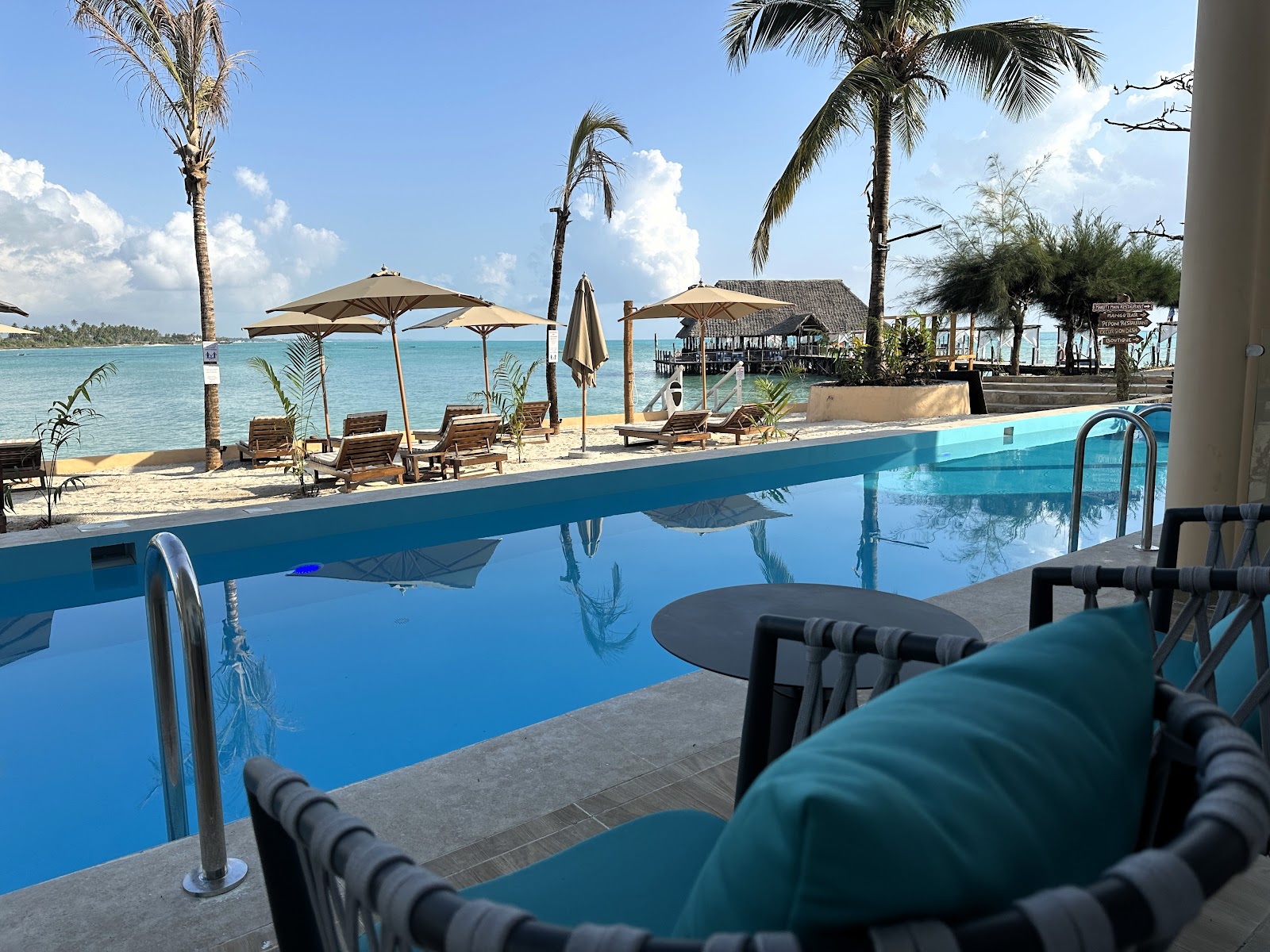 Resort pool with lounge chairs overlooking turquoise lagoon, palm trees, thatched beach structures, and umbrellas on sandy shore.