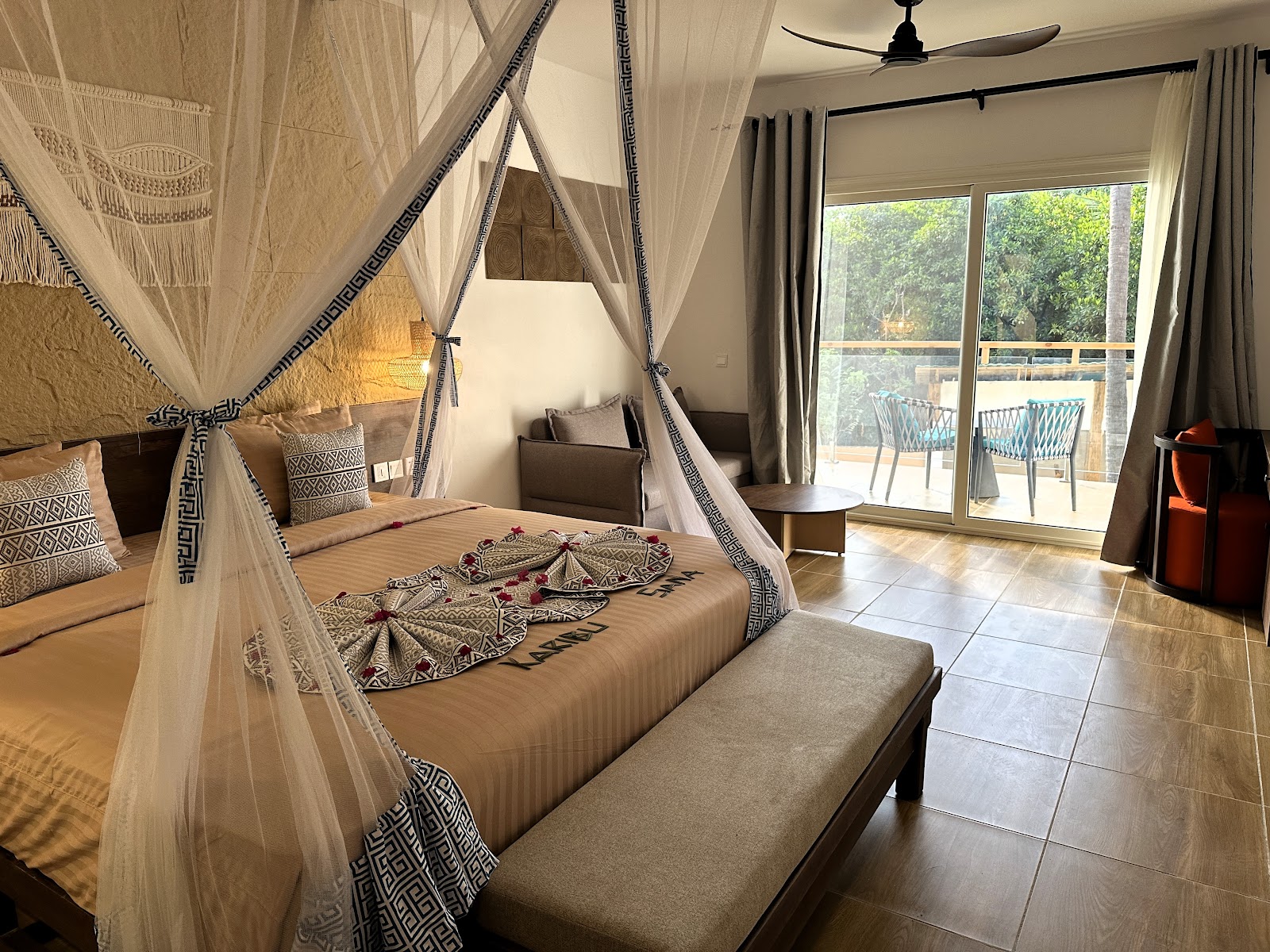 Spacious resort suite with a four-poster canopy bed draped in sheer curtains, tropical decor, and glass doors opening to a balcony overlooking greenery.