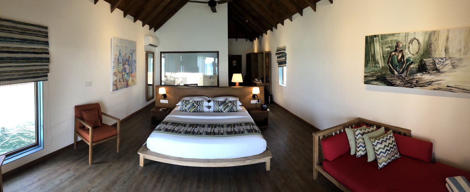 Overwater bungalow interior at Reethi Faru Resort with round bed, dark wood ceiling, red seating, and ocean view through glass doors.