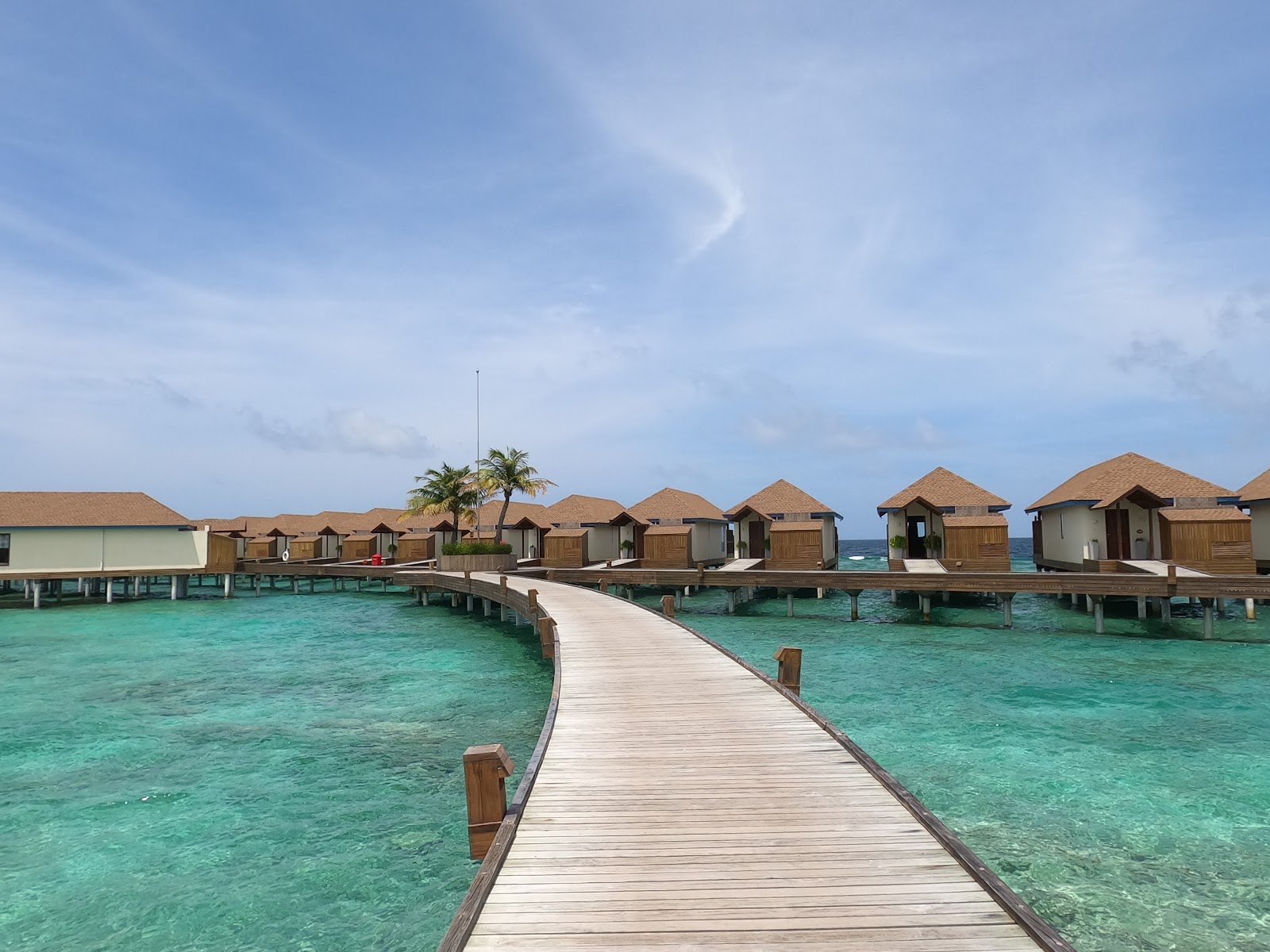 Wooden boardwalk leading to thatched-roof overwater bungalows at Reethi Faru Resort, with turquoise lagoon and palm trees.