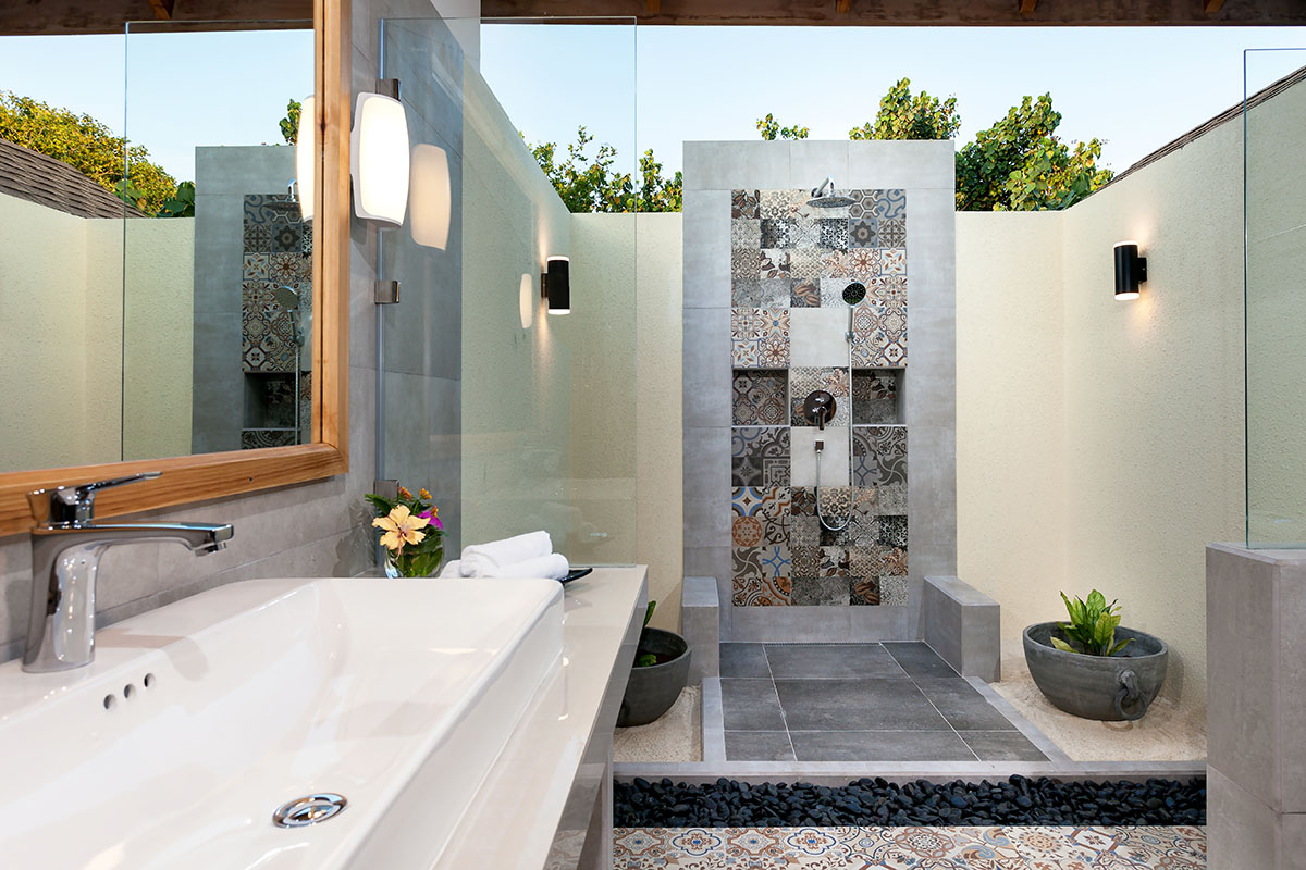 Modern bathroom at Reethi Faru Resort with walk-in shower, white vanity sink, and patterned tile work overlooking tropical grounds.