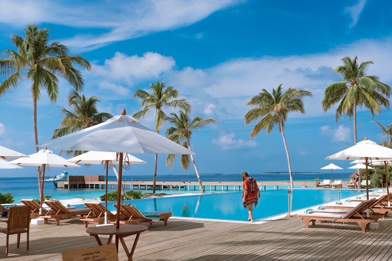 Woman walks past wooden deck with loungers and white umbrellas overlooking turquoise infinity pool and ocean at Reethi Faru Resort.