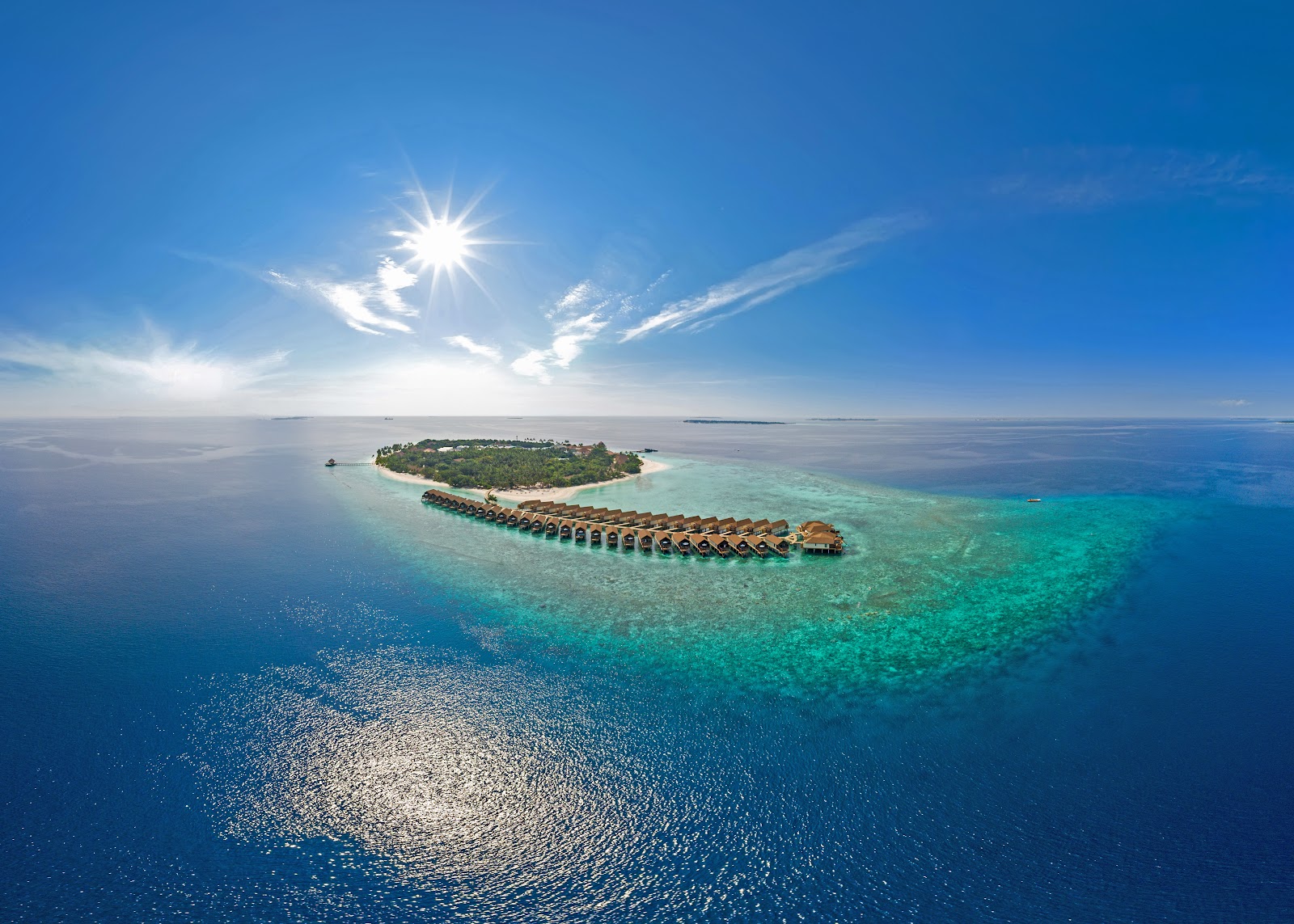 Aerial view of Reethi Faru Resort on a tropical island with overwater bungalows extending into turquoise lagoon waters under bright sunshine.