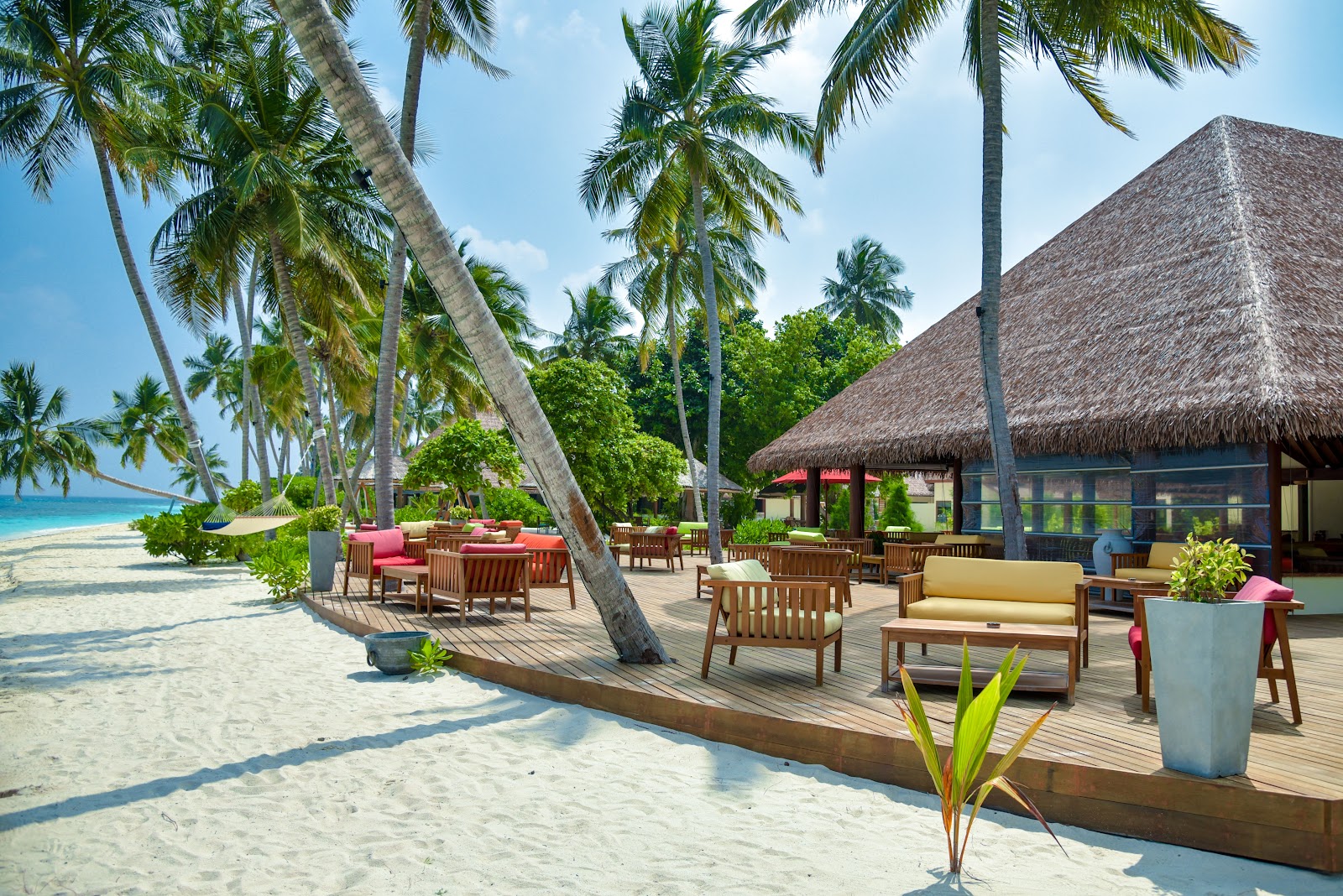 Wooden deck with lounge chairs and dining tables extends from white sand beach under tall palm trees, with thatched-roof bungalows and turquoise ocean visible.