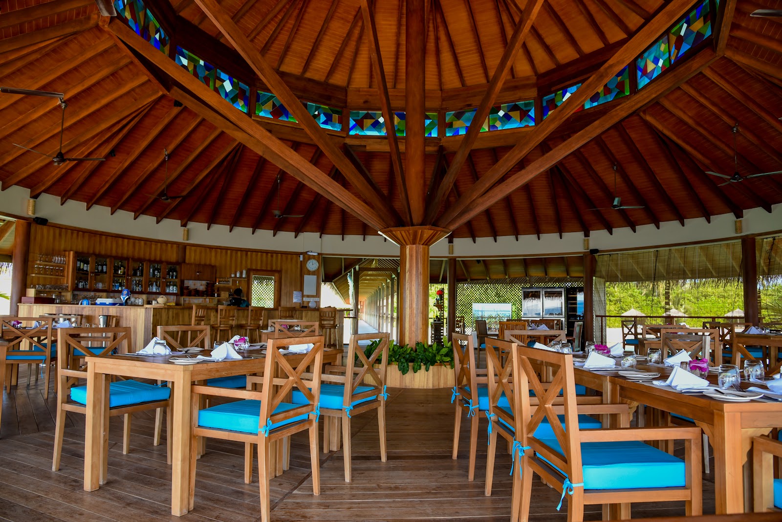 Wooden dining pavilion with stained-glass windows features tables set with blue chair cushions and place settings at Reethi Faru Resort.