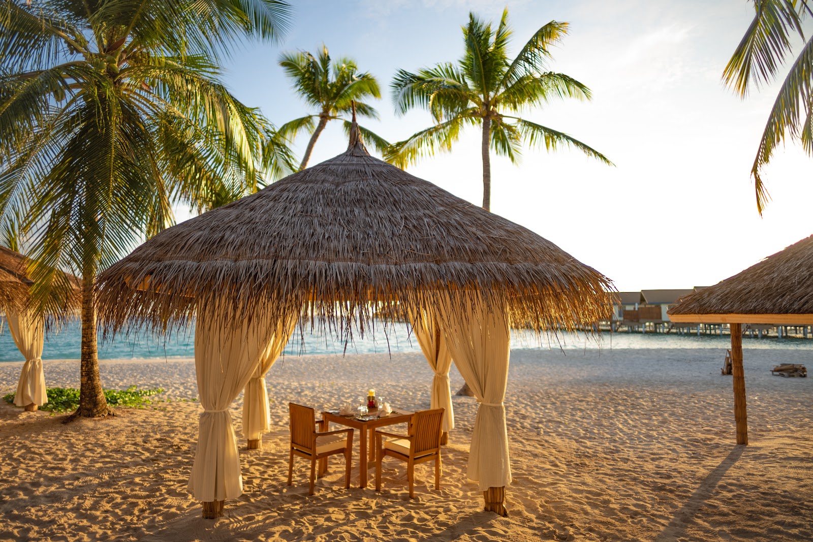Thatched cabana with cream curtains and wooden dining table on sandy beach at Reethi Faru Resort, palm trees and ocean beyond.