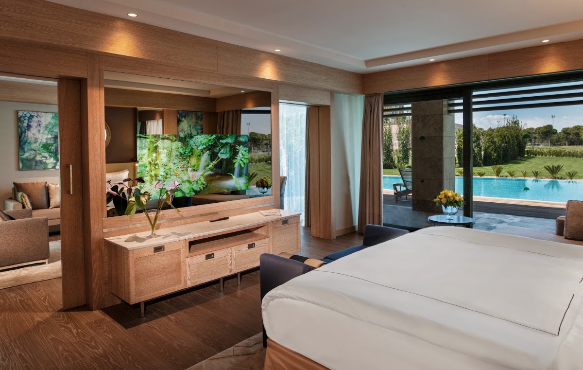 Resort suite with wooden furnishings, white bed, and floor-to-ceiling windows overlooking a pool and landscaped grounds at Regnum Carya Golf & Spa Resort.