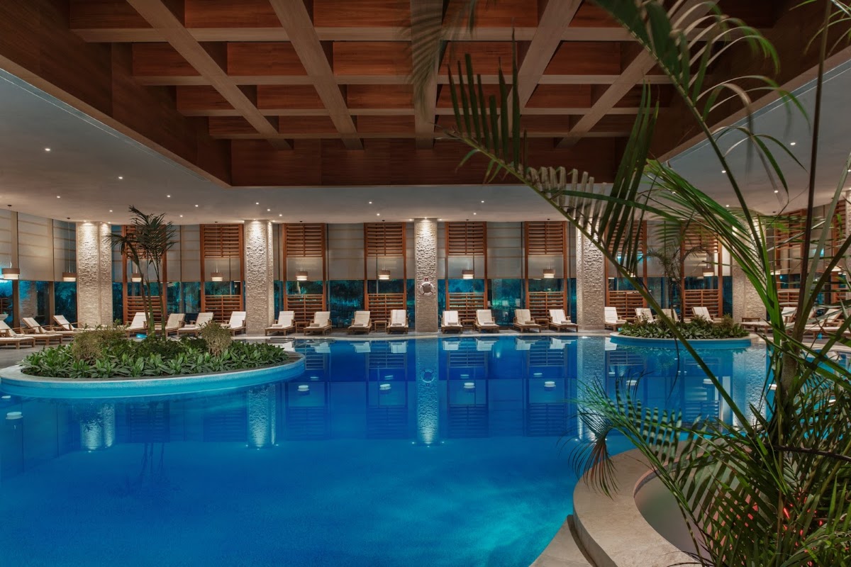 Indoor pool with bright blue water, lounge chairs along the sides, wooden ceiling beams, and tropical plants at Regnum Carya Golf & Spa Resort.