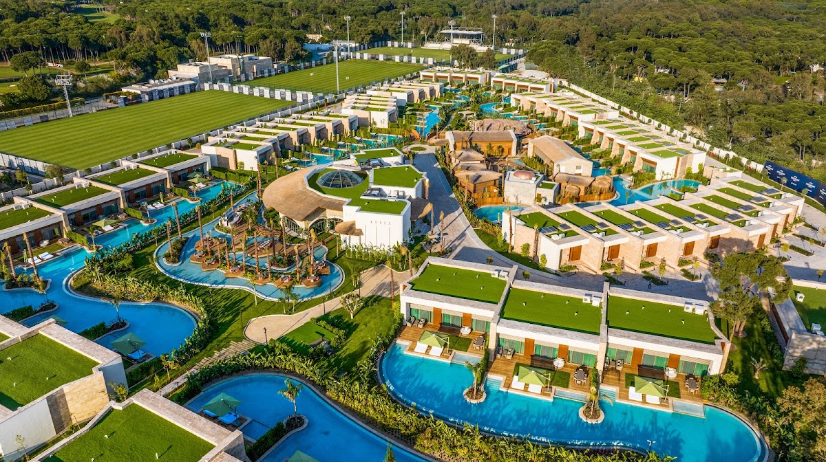 Aerial view of Regnum Carya Golf & Spa Resort showing beige villas with green roofs, blue pools, water slides, manicured lawns, and surrounding forest.