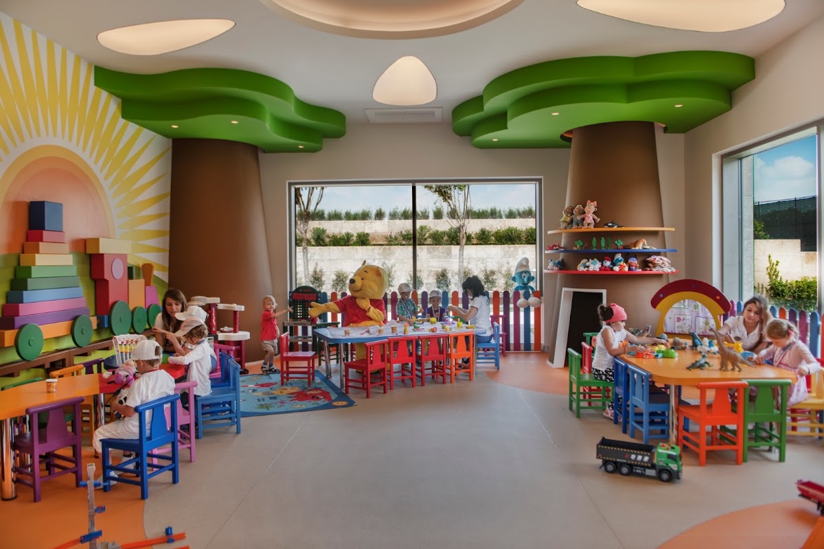 Colorful kids club room with tree-shaped green ceiling elements, rainbow wall art, toy-filled shelves, and children playing at tables with adults supervising.