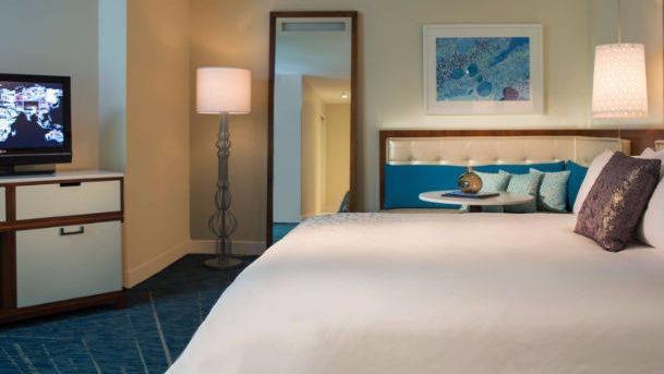 Modern hotel bedroom with white bedding, teal headboard, flat-screen TV, floor lamp, and abstract wall art at Renaissance Wind Creek Aruba Resort.