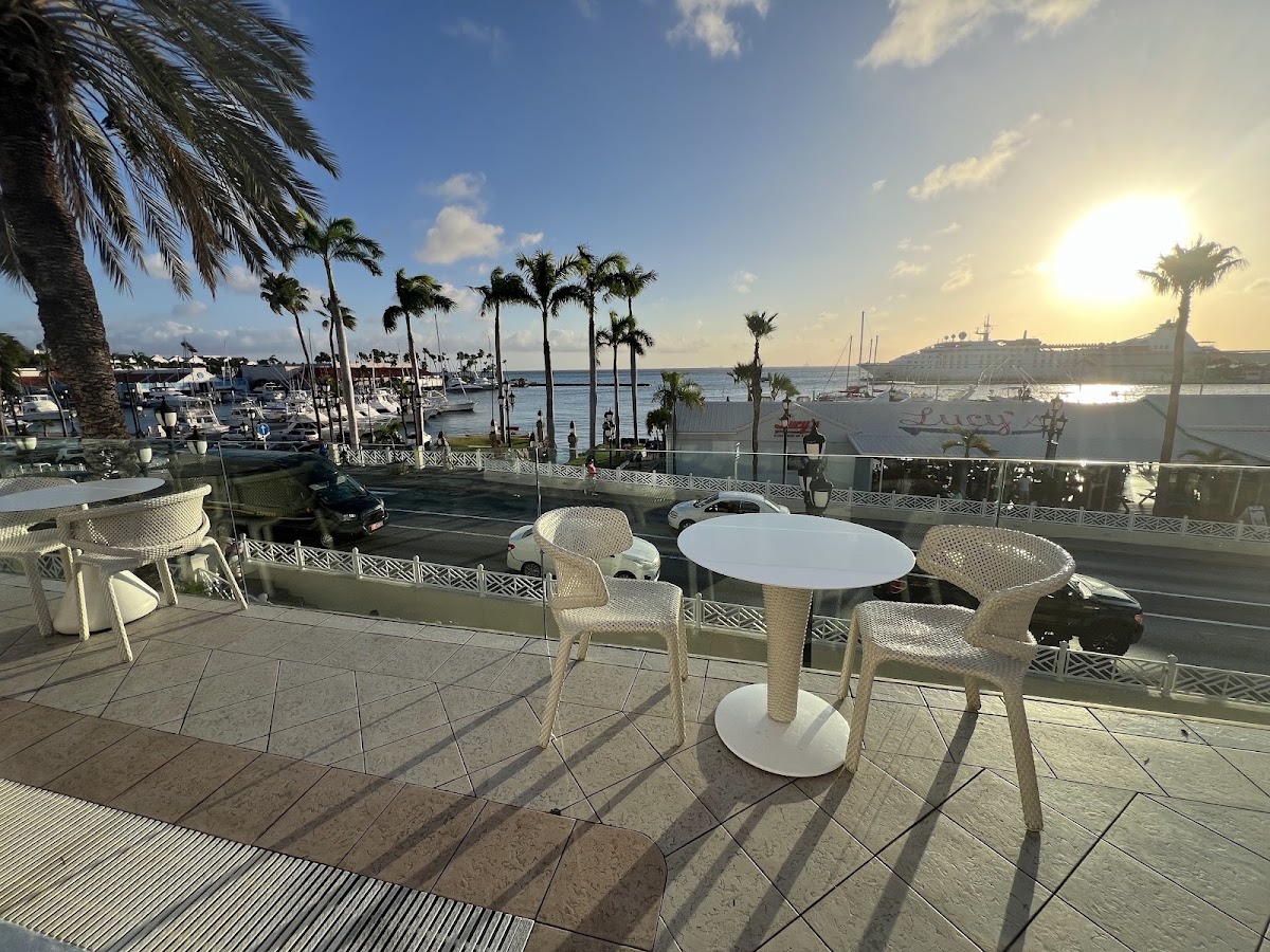 White wicker chairs surround tables on a waterfront terrace at Renaissance Wind Creek Aruba Resort, with palm trees, boats, and the bay visible beyond.