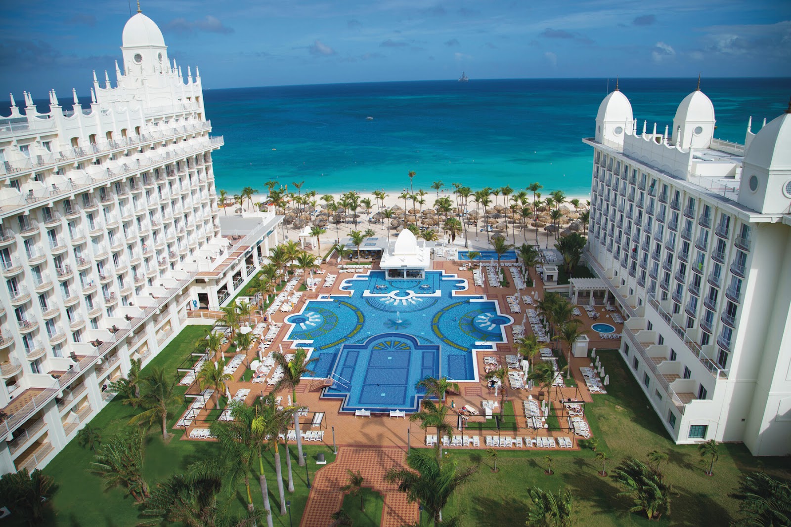 White resort buildings with domed roofs frame a large blue pool complex overlooking a palm-lined beach and turquoise ocean.