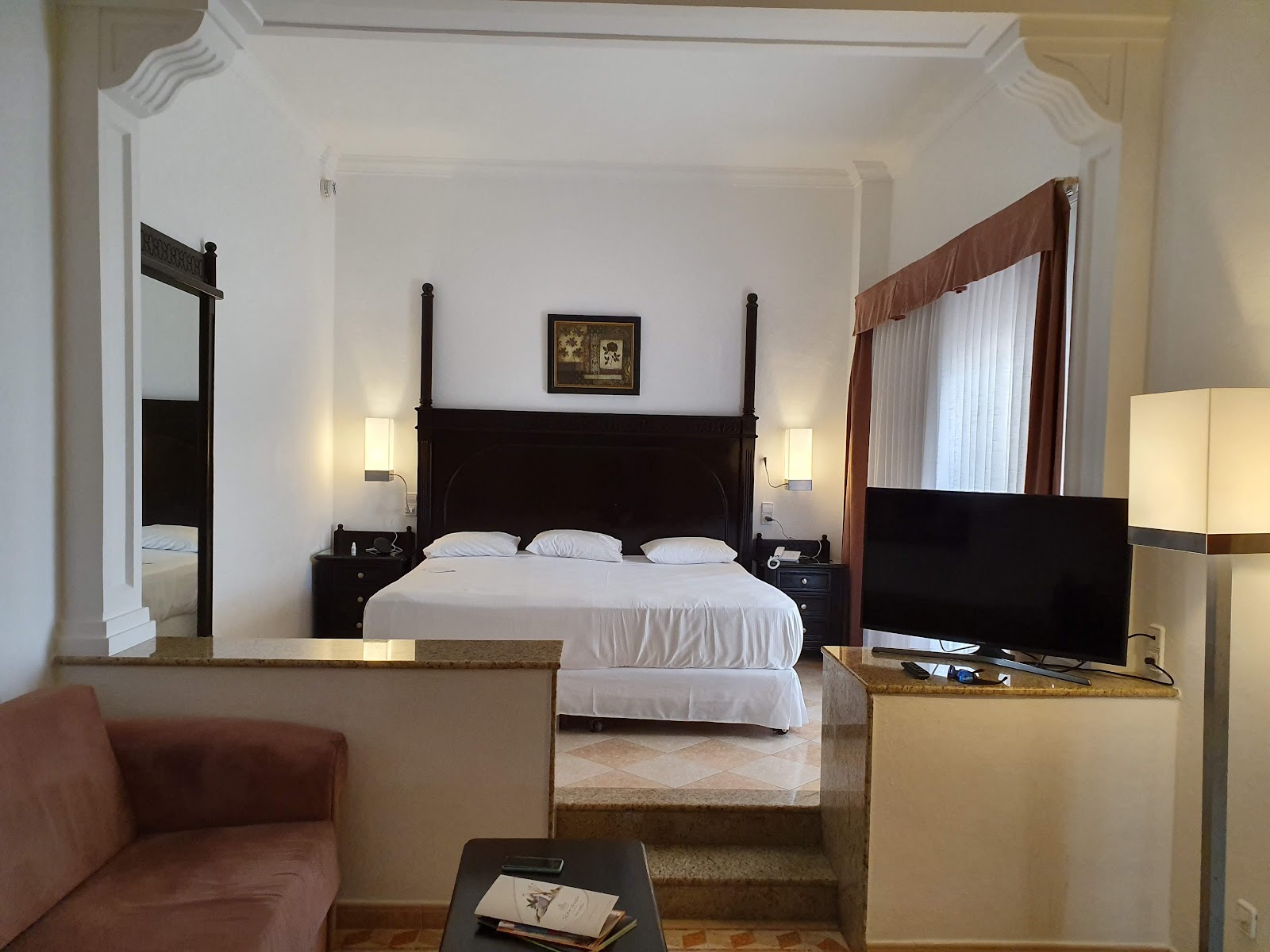 Spacious resort bedroom with four-poster bed, white linens, dark wood furniture, sitting area with burgundy sofa, and flat-screen TV.