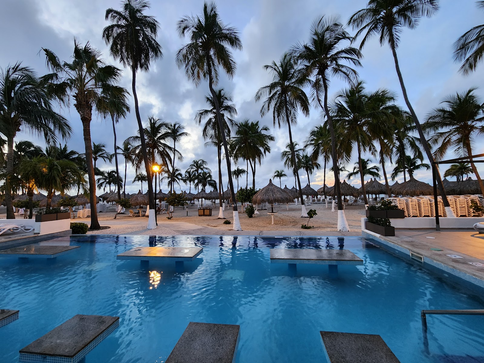 Resort pool with swim-up loungers overlooks palm-lined beach at Hotel Riu Palace Aruba at dusk.