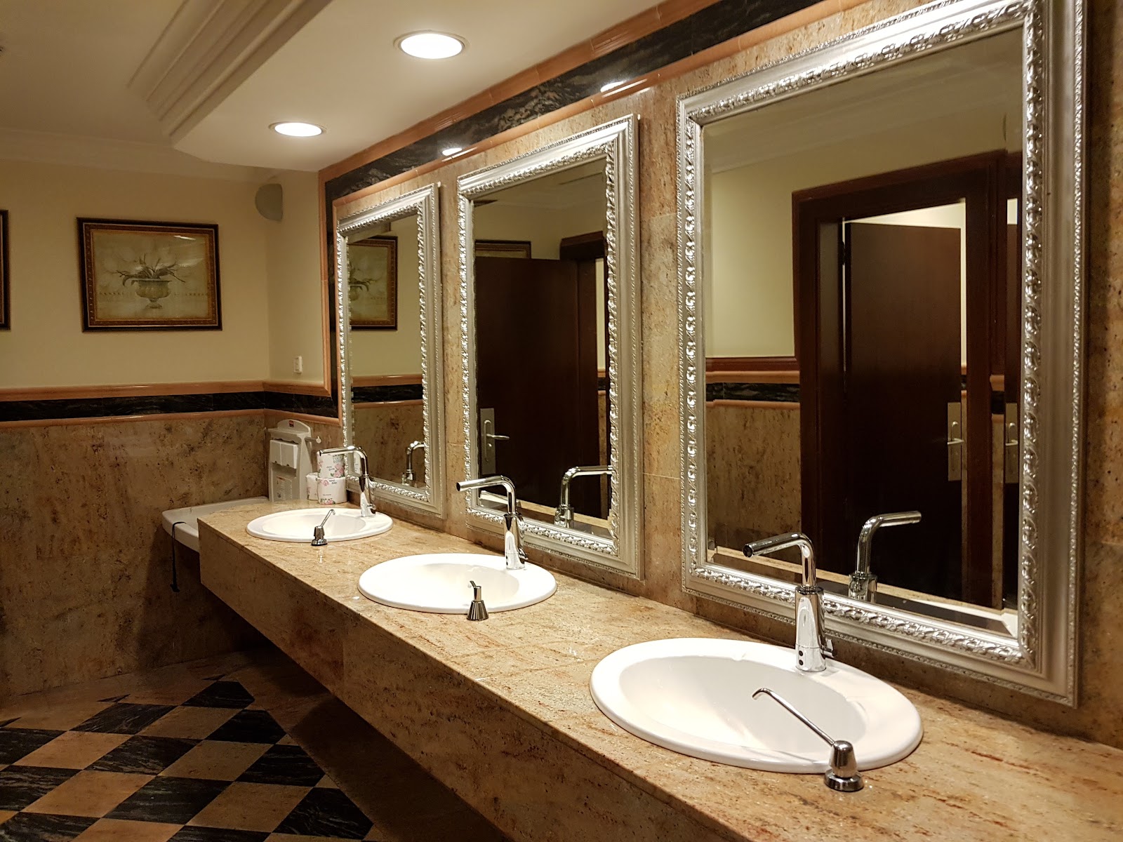Resort bathroom with three white sinks set into a beige granite countertop, ornate framed mirrors, and a checkered tile floor.