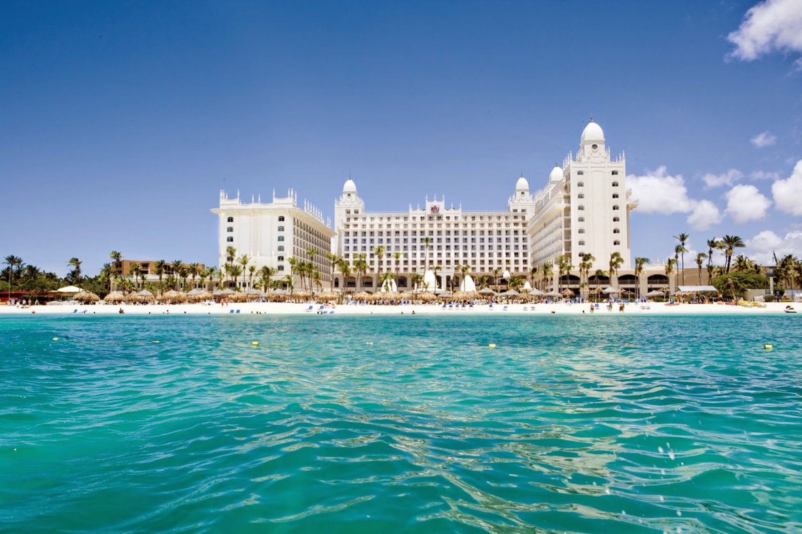Large white resort with distinctive domed towers seen from turquoise ocean with crowded sandy beach and palm trees under blue sky.
