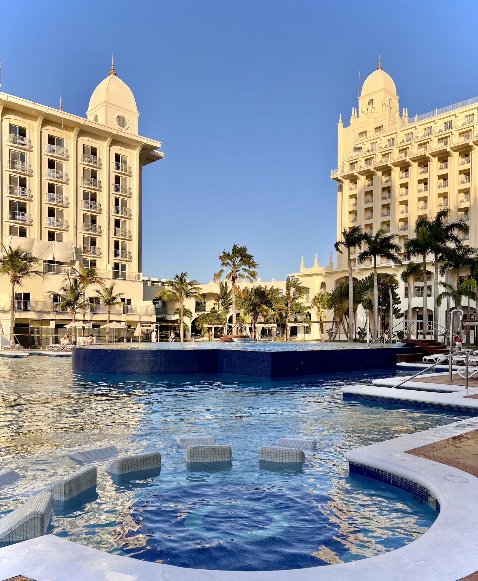 Cream-colored hotel towers with domed roofs overlook multiple swimming pools and palm trees under clear blue sky at Hotel Riu Palace Aruba.