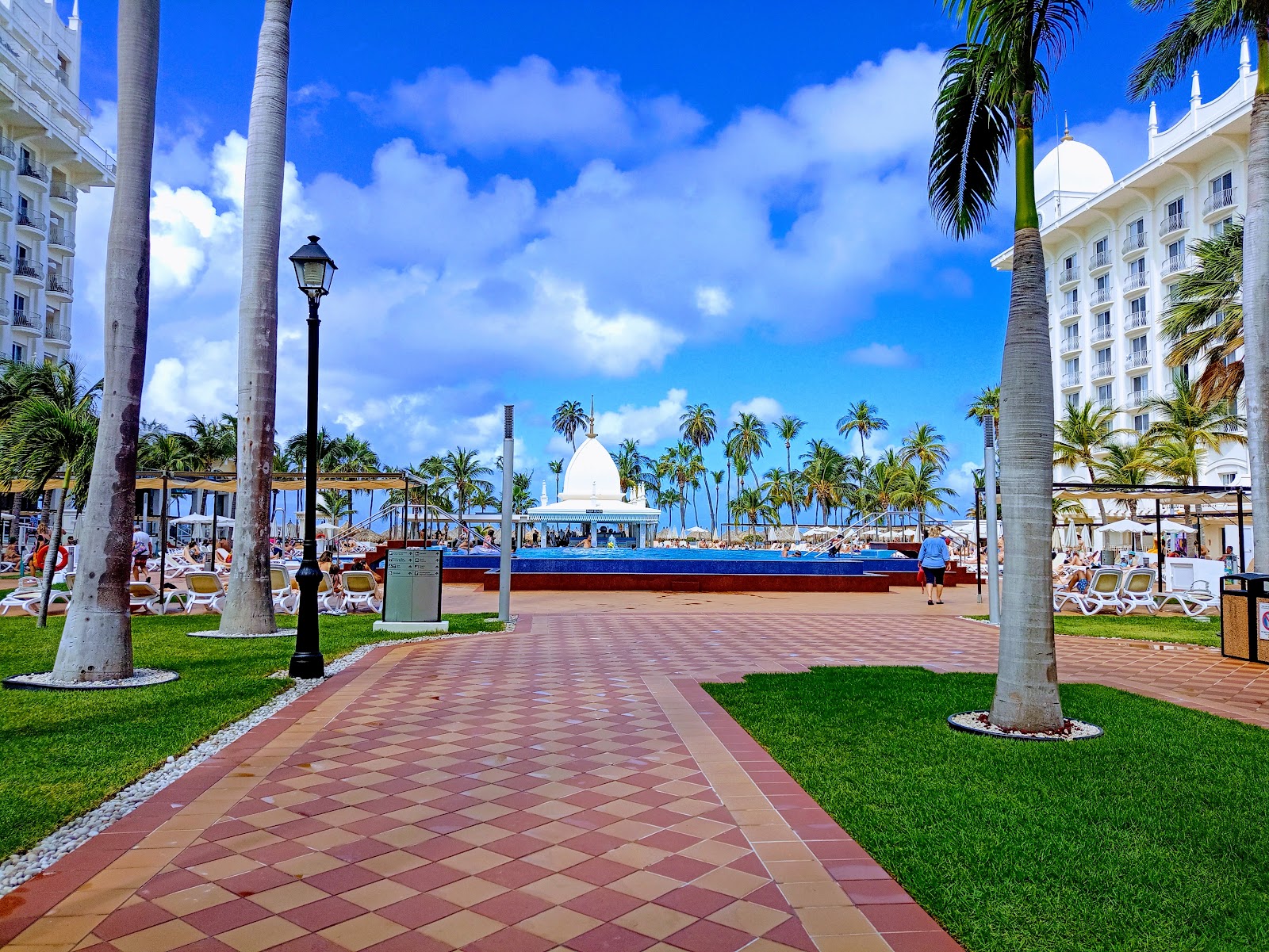 Resort grounds with brick pathway, palm trees, and white hotel buildings flanking a beach with white gazebo and blue cruise ship in distance.