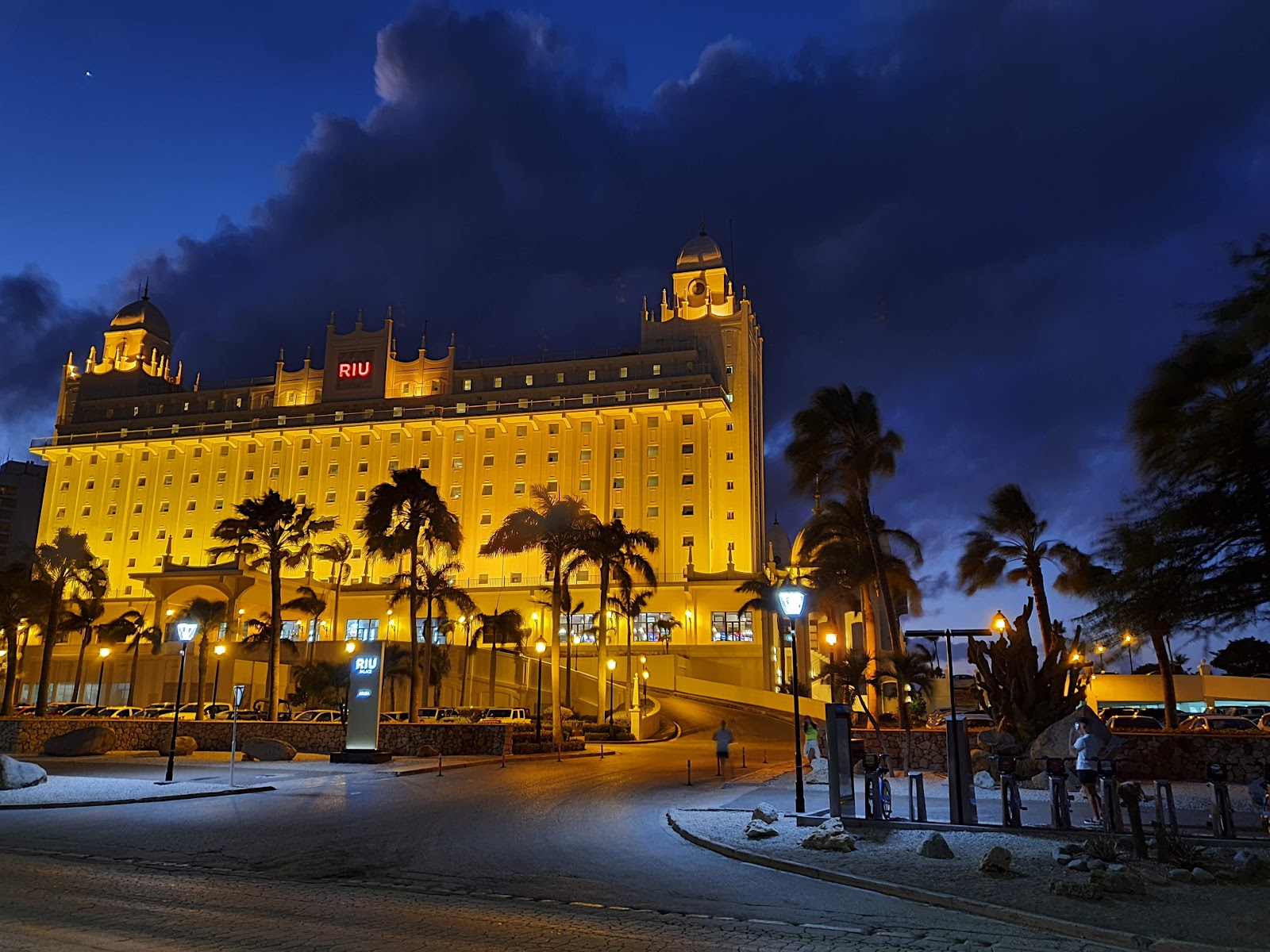 Hotel Riu Palace Aruba illuminated at dusk with palm trees lining the beachfront plaza and dramatic blue sky above.