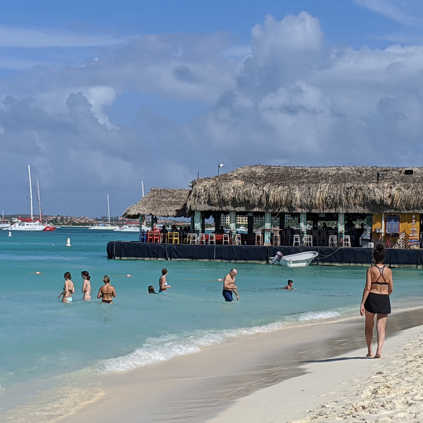 Guests swim and wade in turquoise water off a sandy beach with a thatched-roof beach bar and moored sailboats in the background.