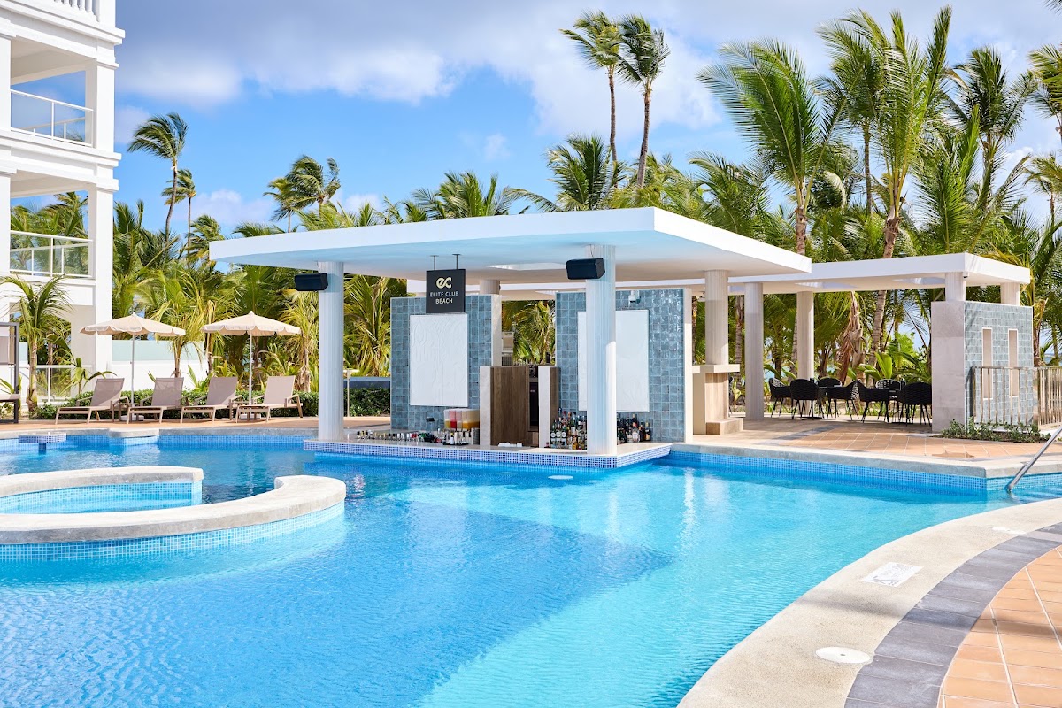 Blue lagoon-style pool with white pergola bar, palm trees, and white resort building under clear sky.