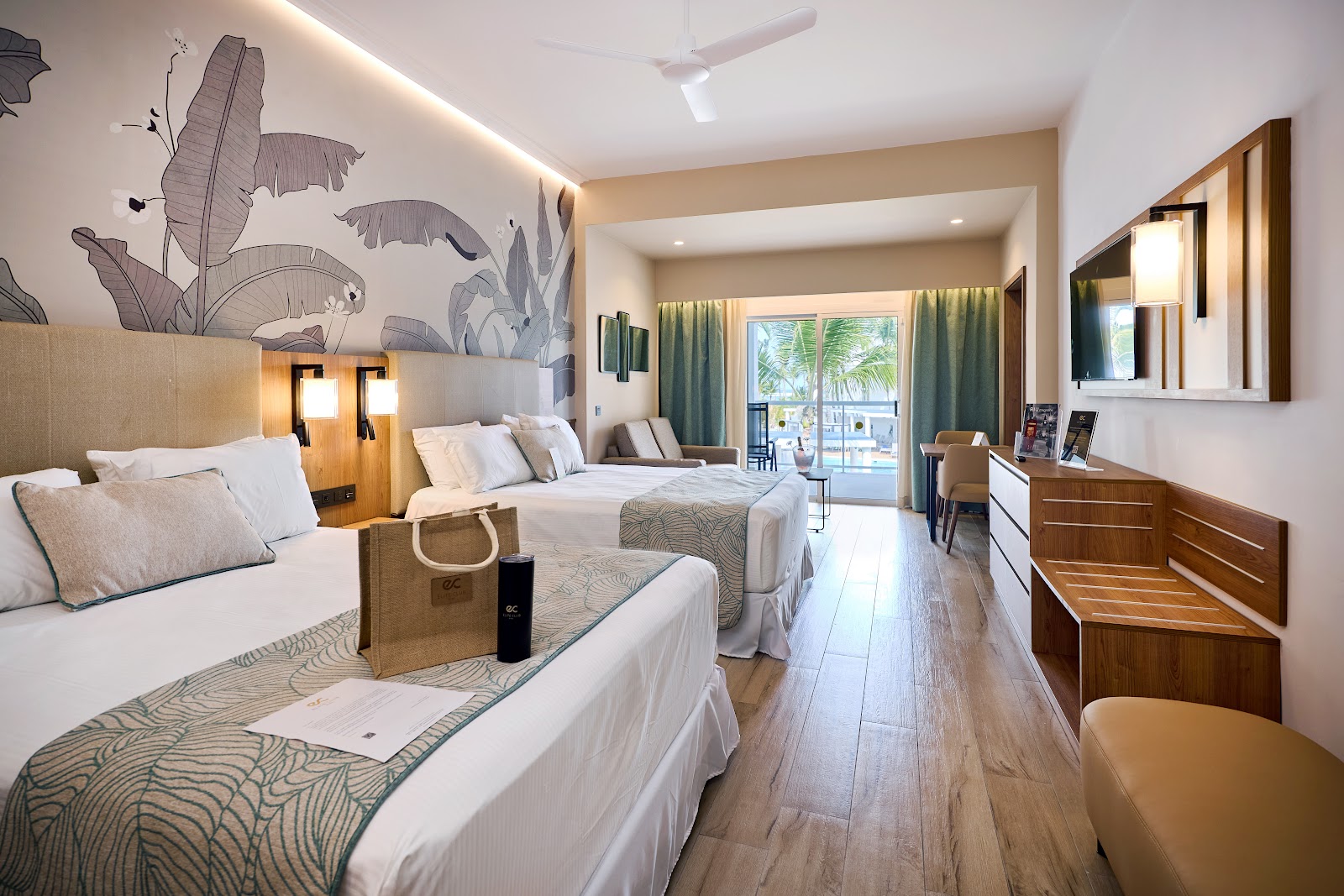 Resort room with two beds, tropical leaf wall mural, wooden furnishings, and ocean view through glass doors to the balcony.