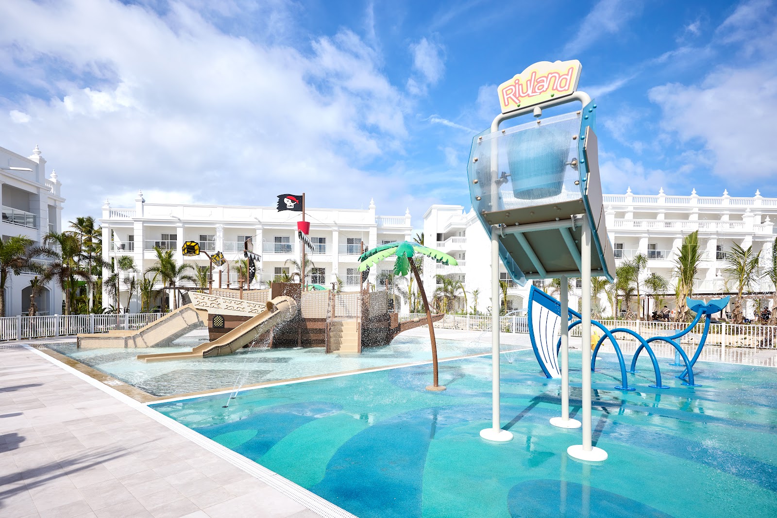 Splash pad and shallow pool at Riu Palace Bavaro with blue water bucket dumper, slide, and white resort building in background.