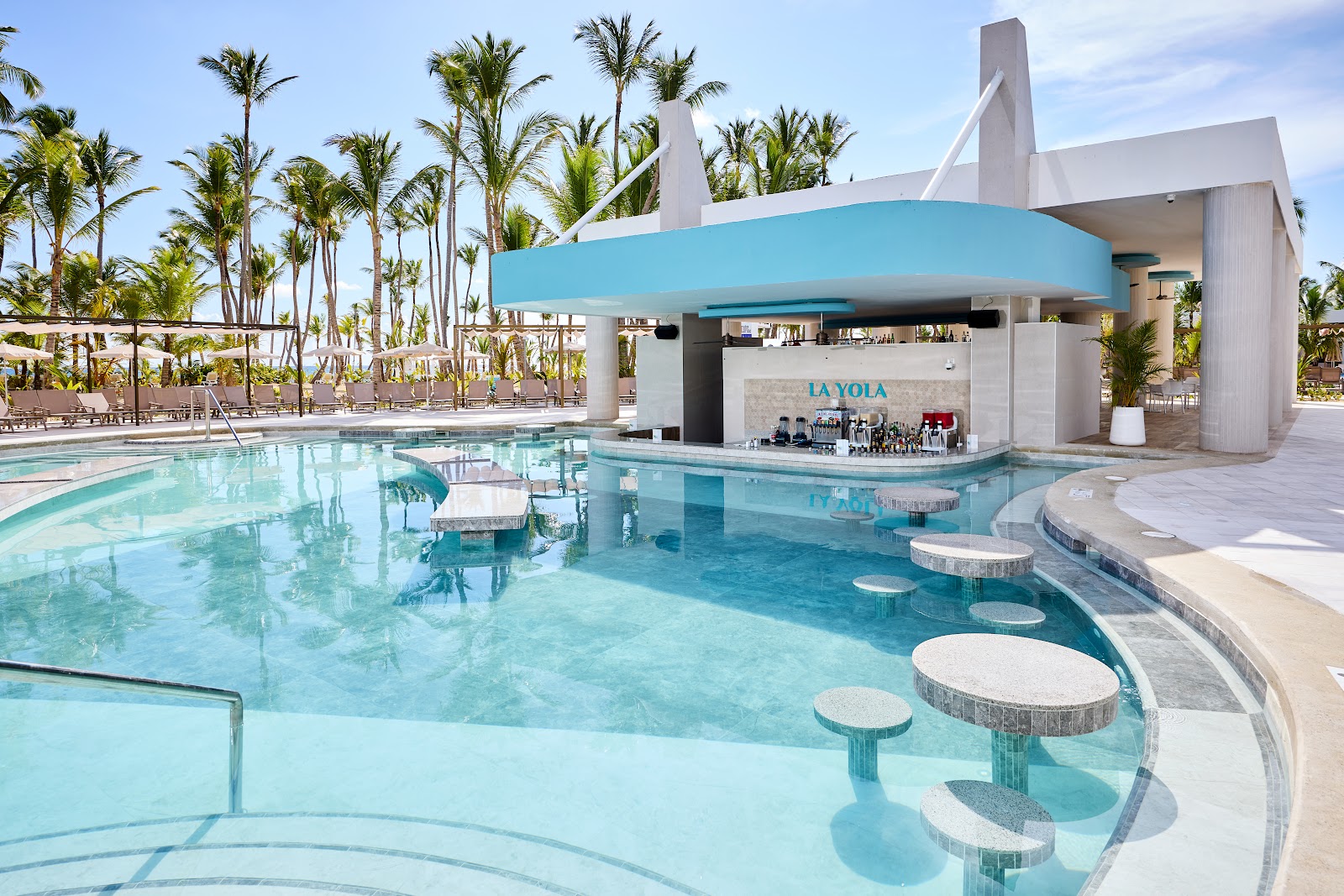 Modern pool bar with turquoise water and submerged seating at La Voya, surrounded by palm trees and lounge chairs under clear sky.