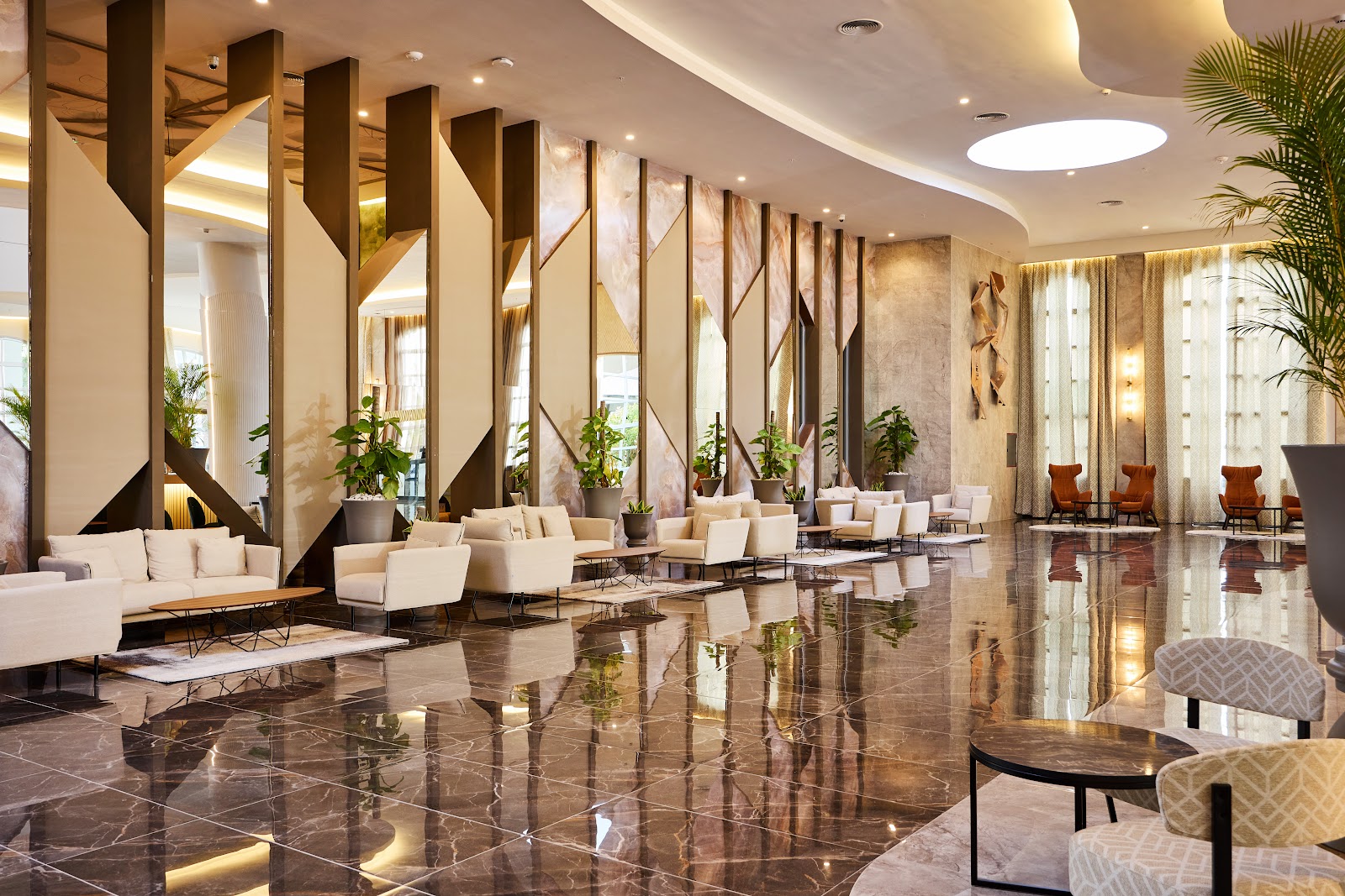Modern lobby with polished dark marble floors, cream seating areas, tall wooden dividers, potted plants, and curved ceiling with skylight at Riu Palace Bavaro.