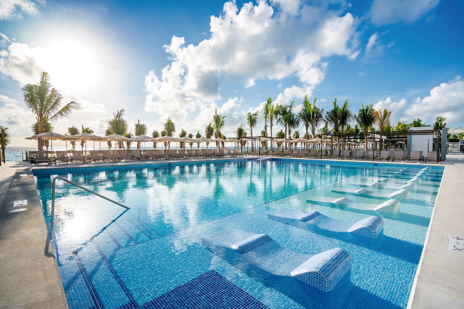 Large pool with submerged loungers, palm trees, and thatched umbrellas under blue sky at Riu Palace Kukulkan.