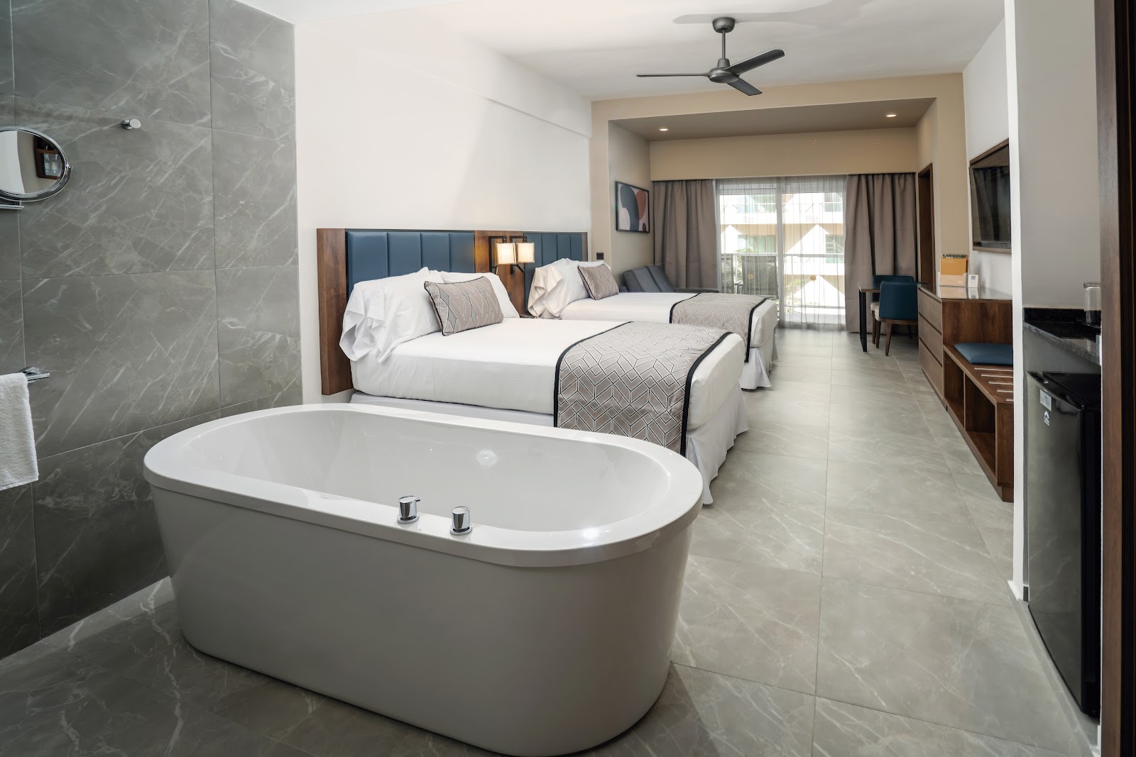 Freestanding white bathtub in foreground with two queen beds, blue headboards, and glass shower enclosure in spacious resort suite.