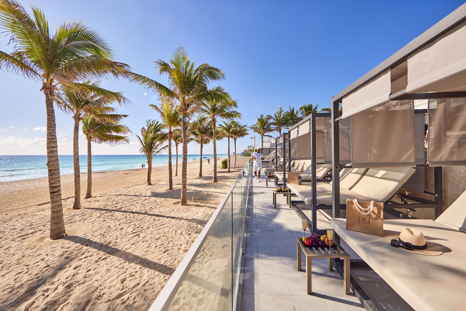 Beachfront cabanas with shade structures line the sandy beach at Riu Palace Kukulkan, with palm trees and turquoise ocean beyond.