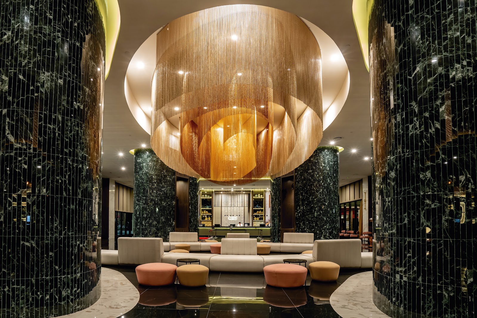 Modern resort lobby with a large spherical wood chandelier, black marble pillars, cream modular seating, and a reception desk at the Riu Palace Kukulkan.
