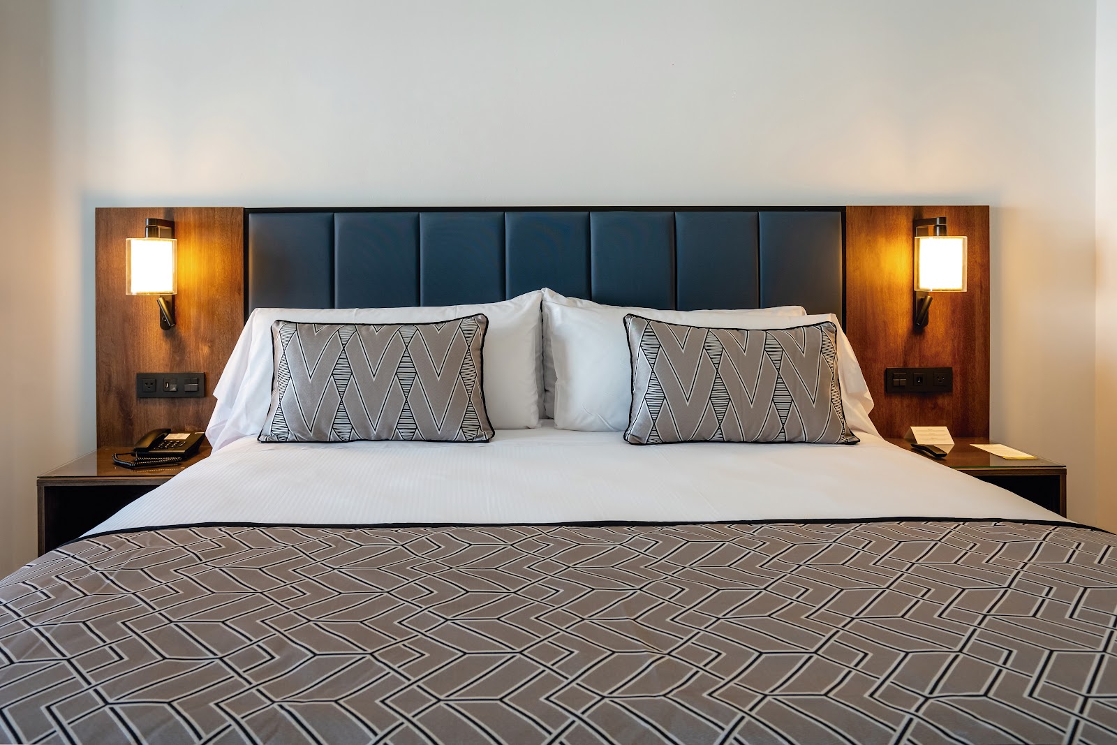 Resort bedroom with king bed, dark blue upholstered headboard, wood-paneled nightstands with warm lighting, and geometric patterned pillows.
