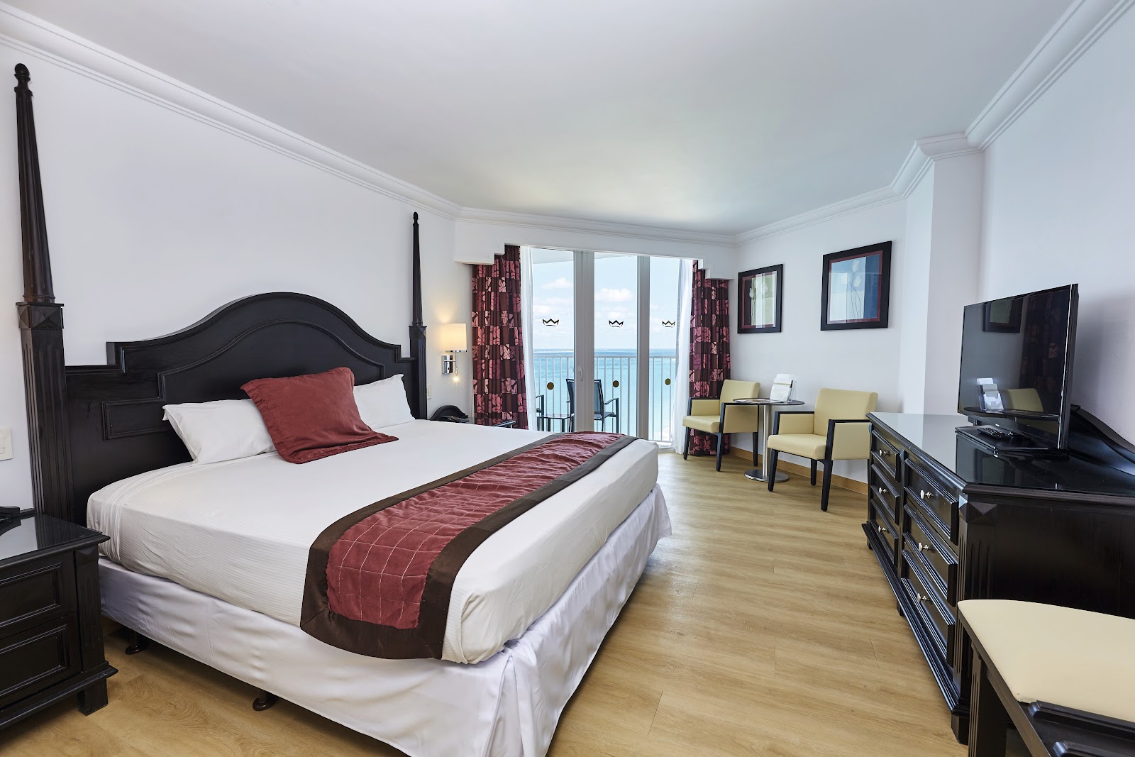Resort guestroom with black four-poster bed, ocean view balcony, sitting area with yellow chairs, and flat-screen TV.